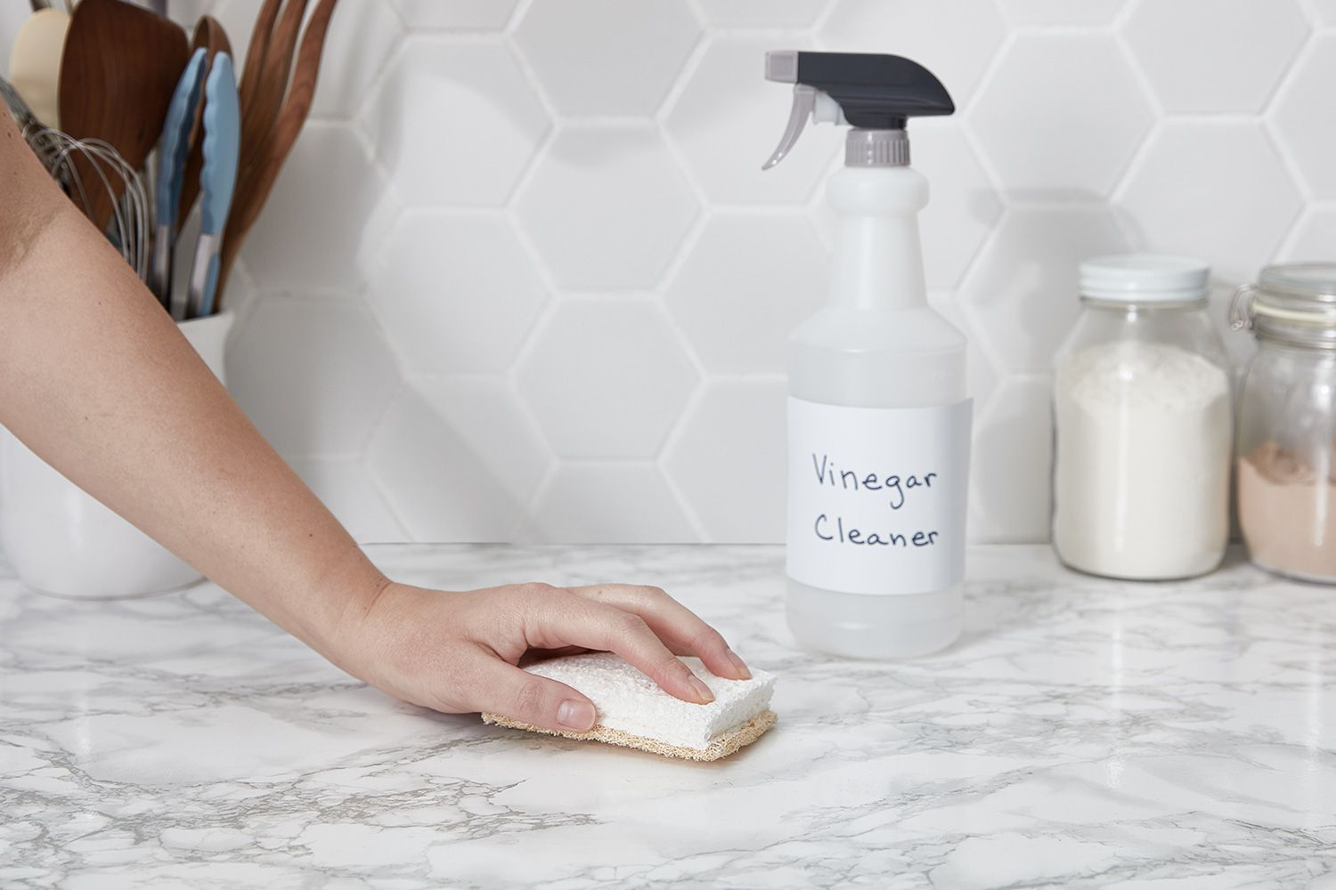A person cleans a marble countertop with a sponge, next to a vinegar cleaner spray bottle and jars in a bright kitchen.