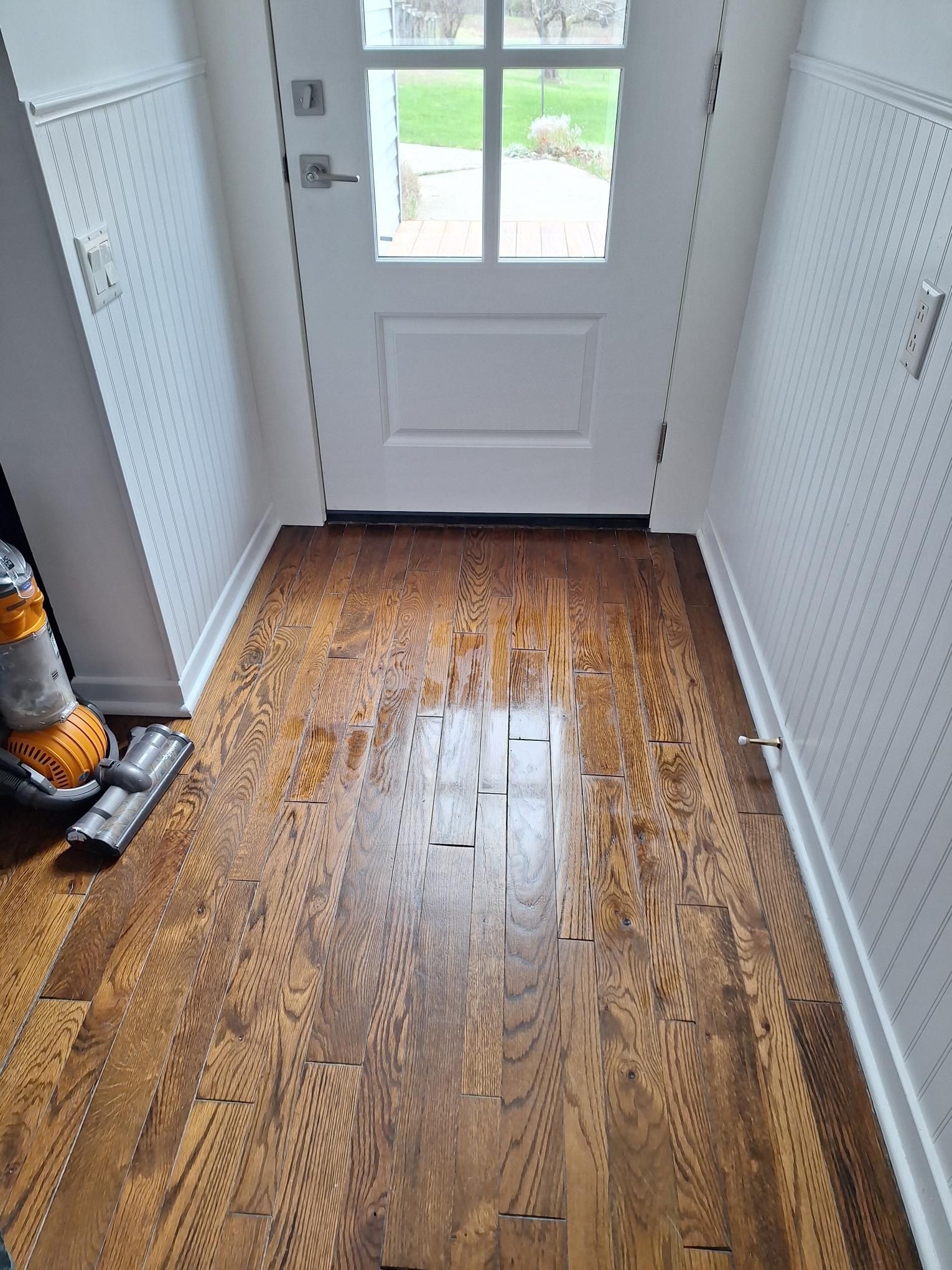An entryway with white beadboard walls and a wooden floor, showing a patch of worn finish near a white door.