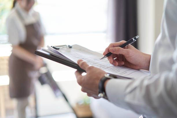 Person with clipboard taking notes, in front of a person vacuuming in a room.