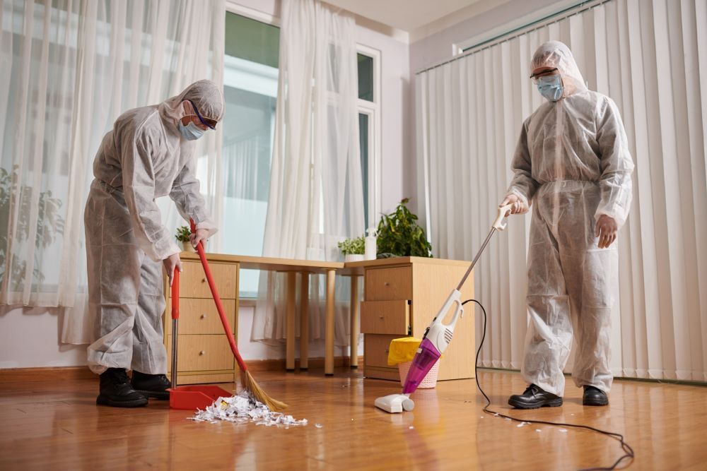 Two people in hazmat suits cleaning a room with a broom, dustpan, and vacuum.