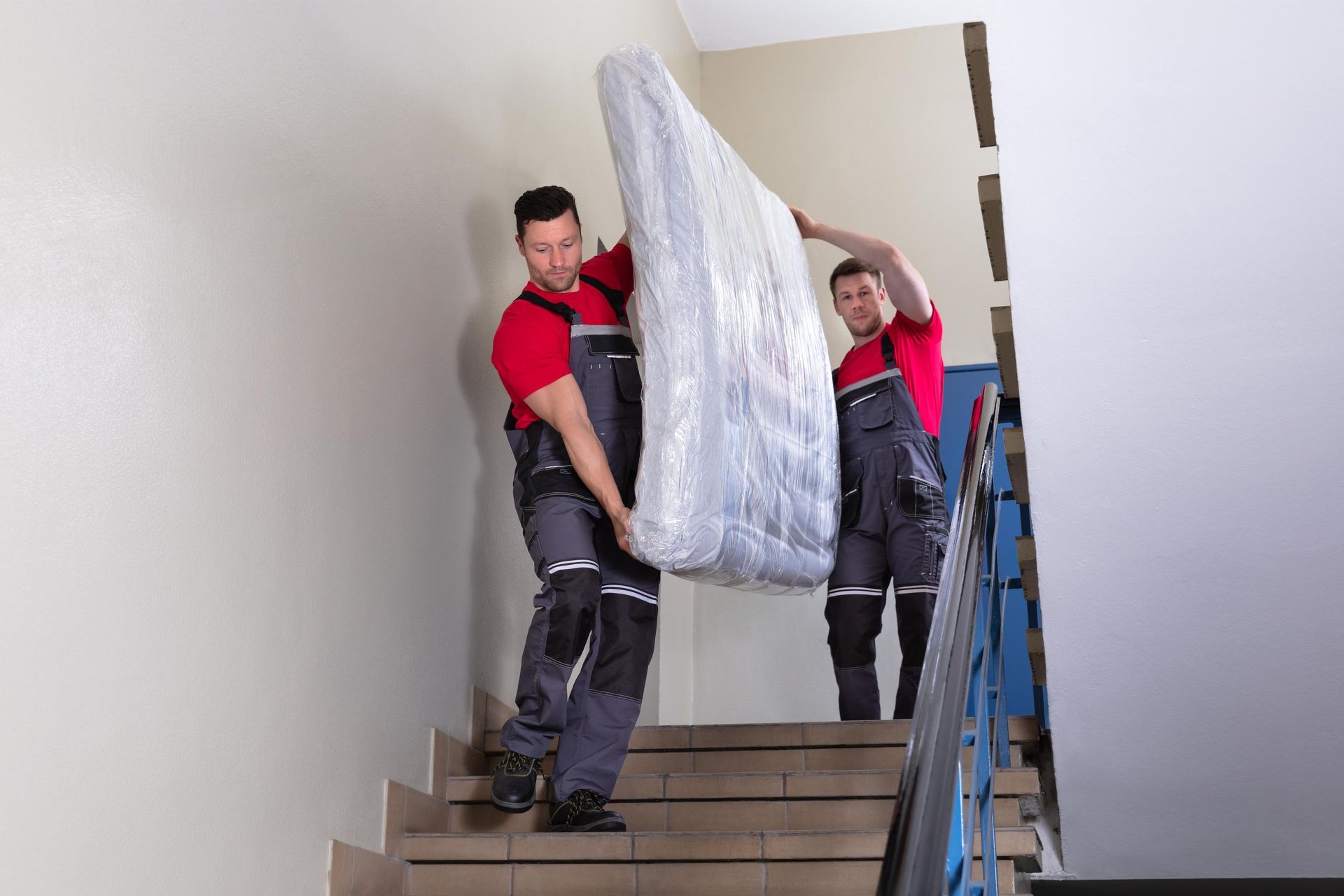 Two movers carrying a wrapped mattress down a staircase.
