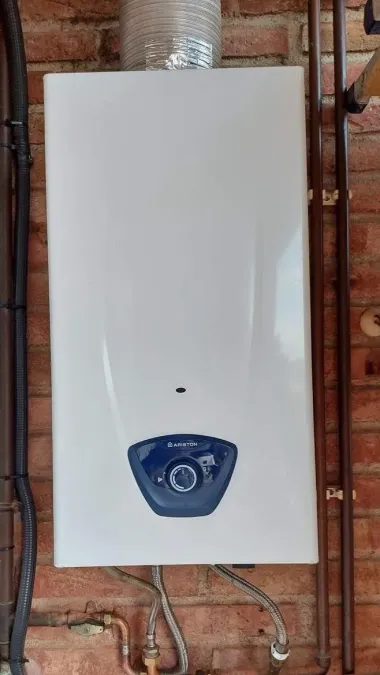 White wall-mounted boiler with a blue control panel, installed against a brick wall with exposed pipes.