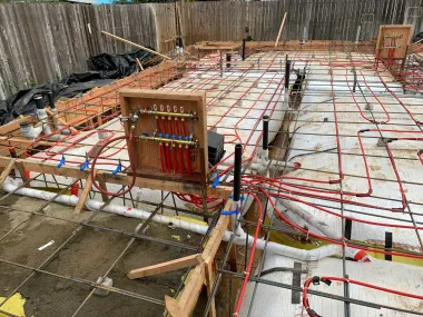 Foundation slab under construction with rebar grid, red tubing, and wooden formwork in a fenced backyard.