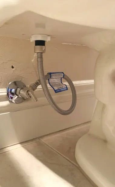 Chrome shut-off valve and flexible hose under a sink, with a blue warning tag in a white cabinet