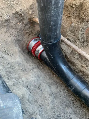 Black pipe joint with red coupling in a dirt trench beside a vertical pipe