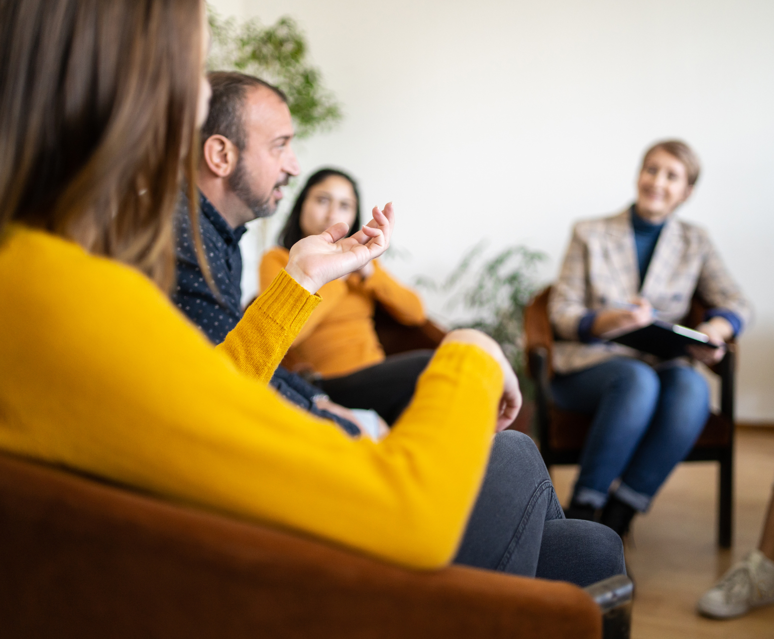 People in therapy session, woman gestures, counselor takes notes, bright room.
