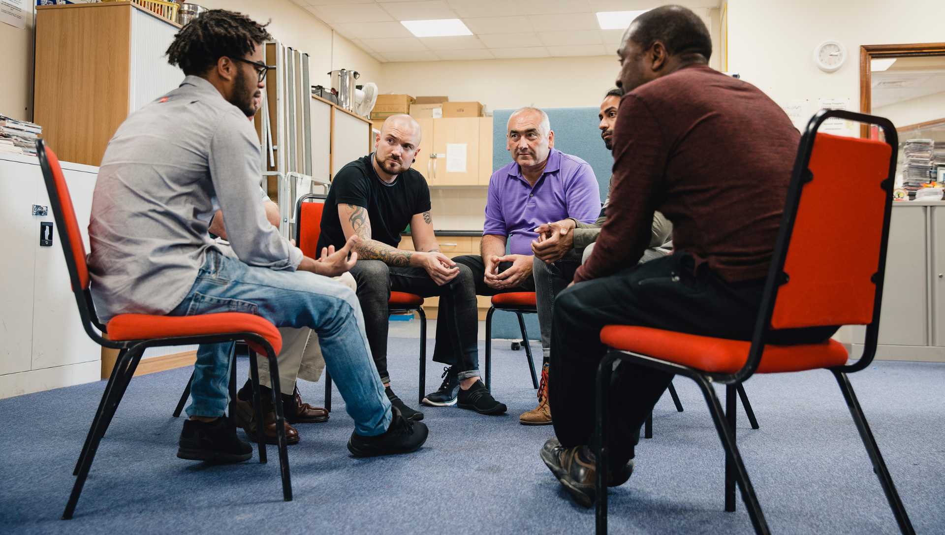 Five people in a circle, engaged in a group discussion. Indoor setting, red chairs, neutral expressions.