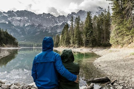 A man and a woman are sitting on the shore of a lake looking at the mountains.