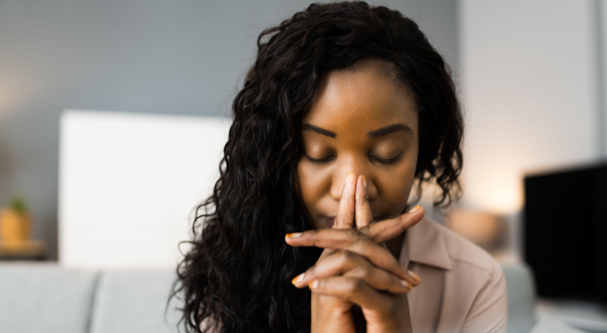 A Black woman with long curly hair, eyes closed, hands clasped in prayer, appears to be thoughtful indoors.