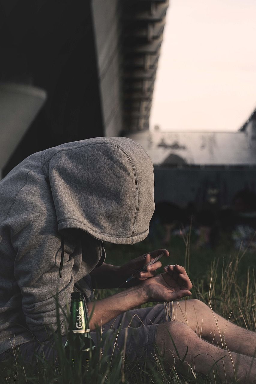 Person in a gray hoodie, slumped over with a needle and beer bottle, outside under a bridge.