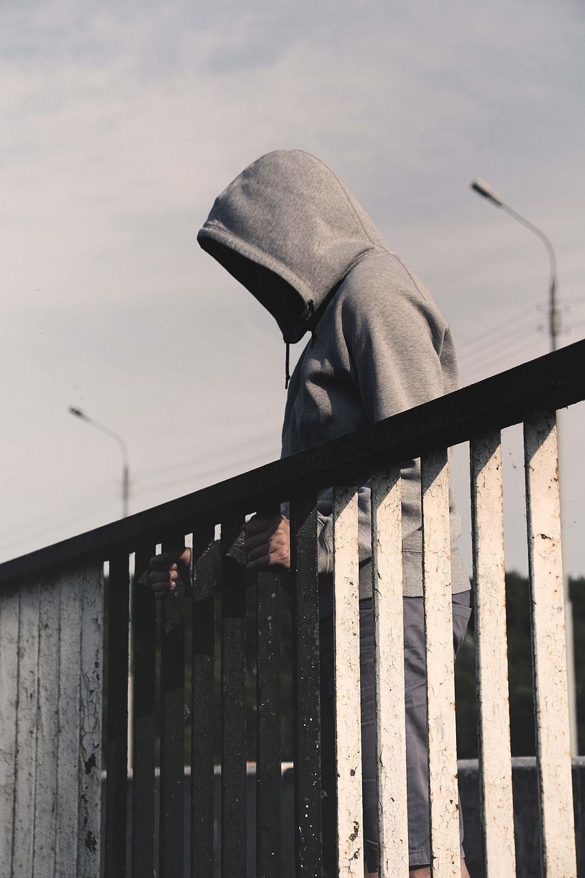 Person in a gray hoodie, head down, stands near a white railing with a cityscape background.