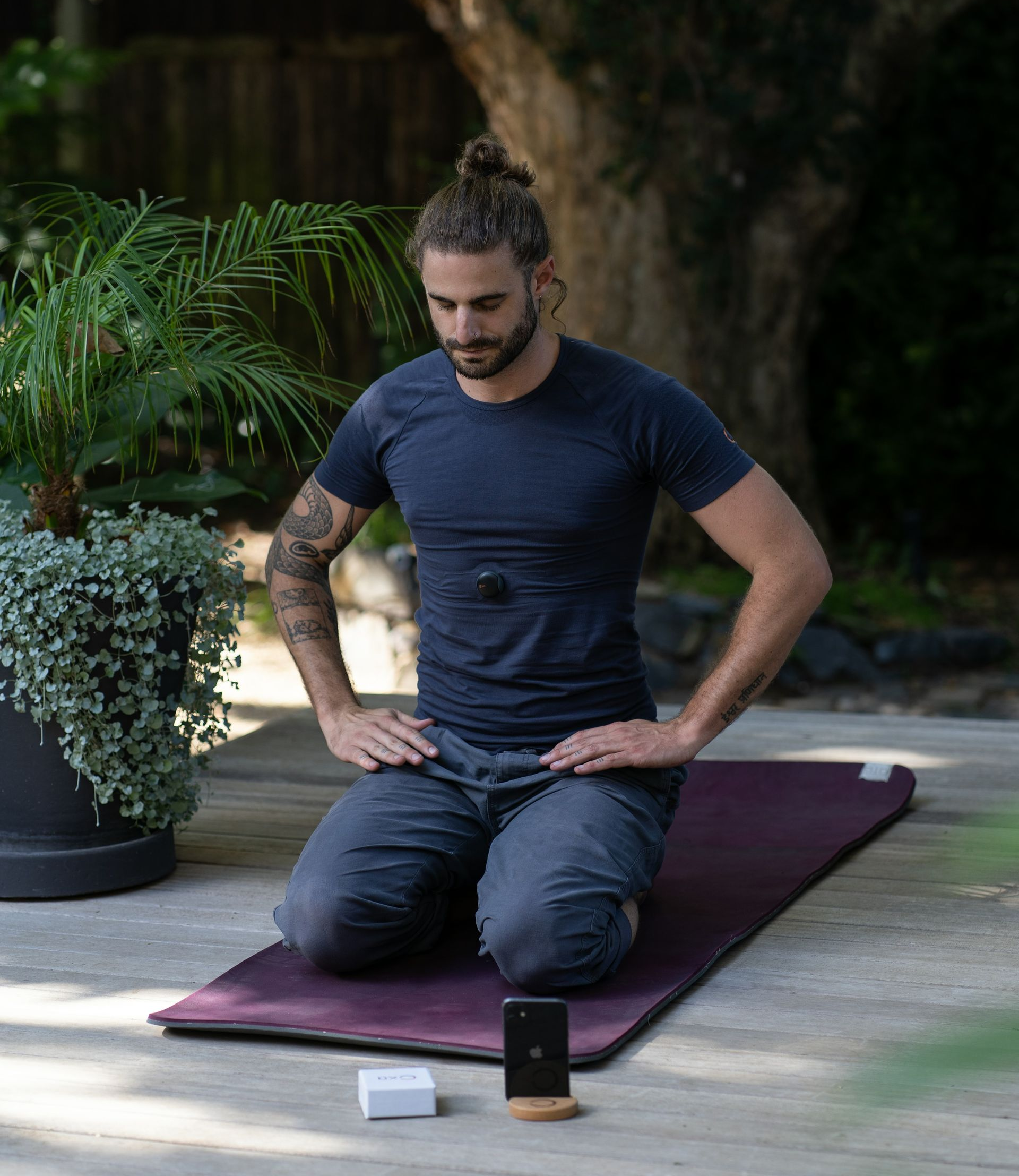 Man meditating on a mat outdoors, hands on knees. Wearing a sensor on his chest. Phone and box nearby.