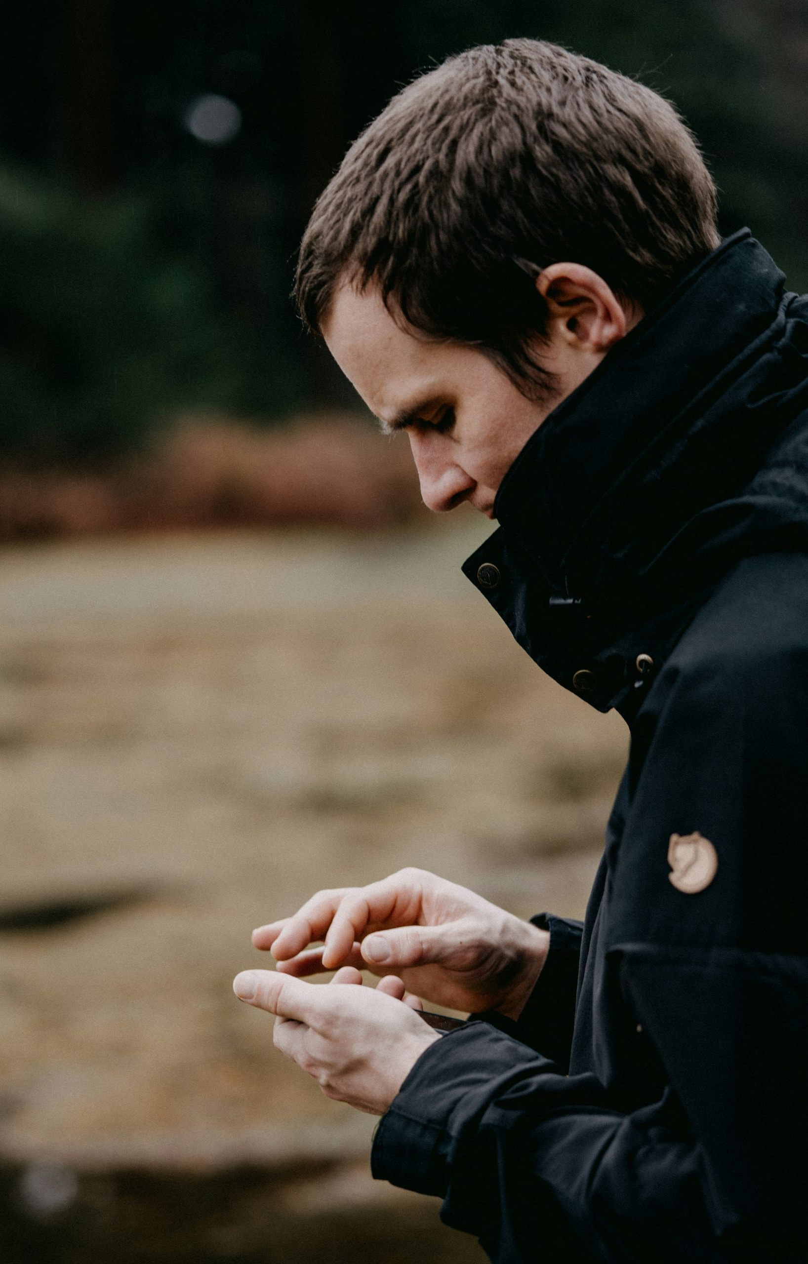Man in dark jacket looking down at his hands, likely holding something, outdoor setting.