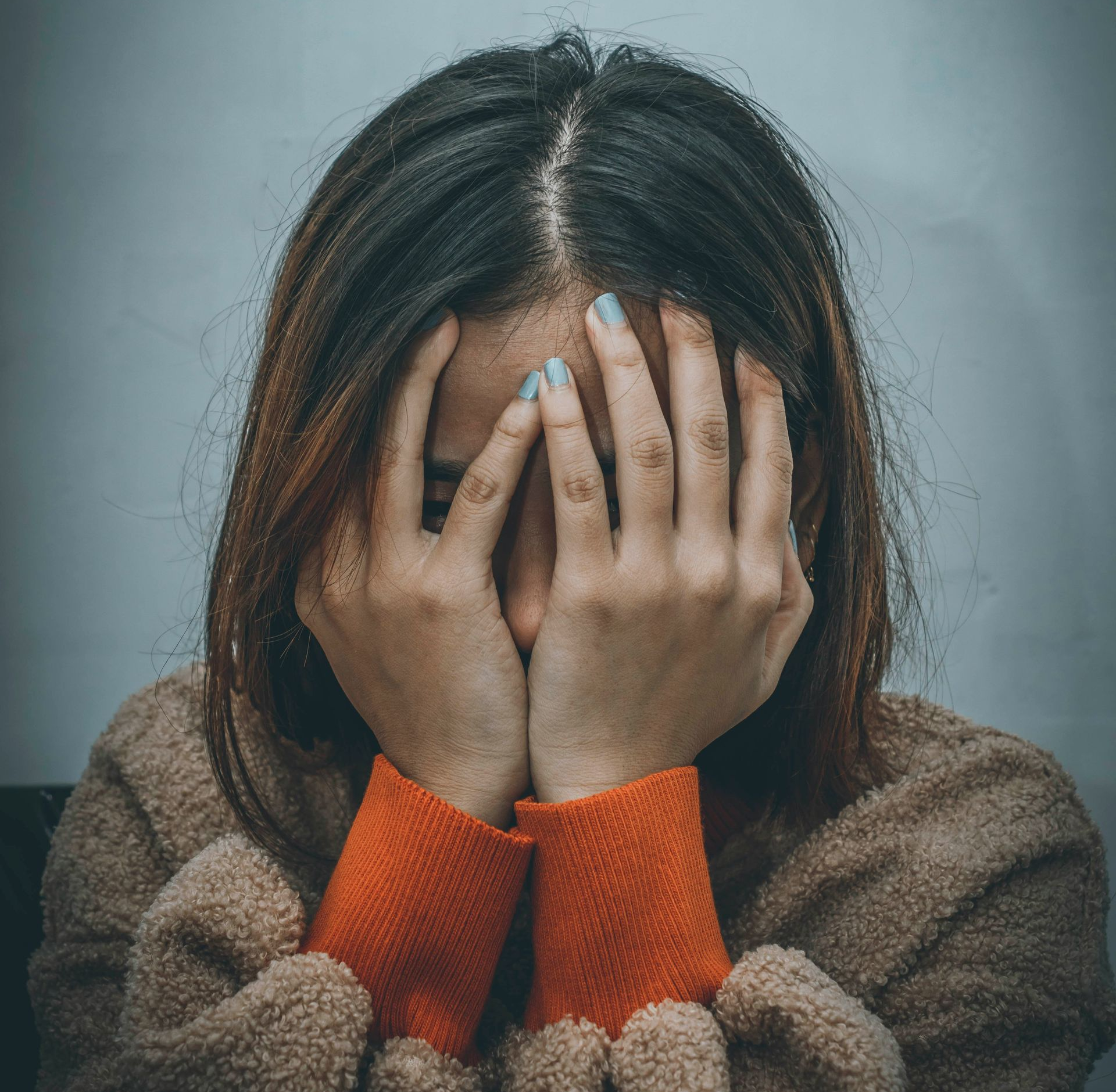 Woman with hands covering face, appearing distressed. Wearing a brown sweater with orange cuffs; dim lighting.