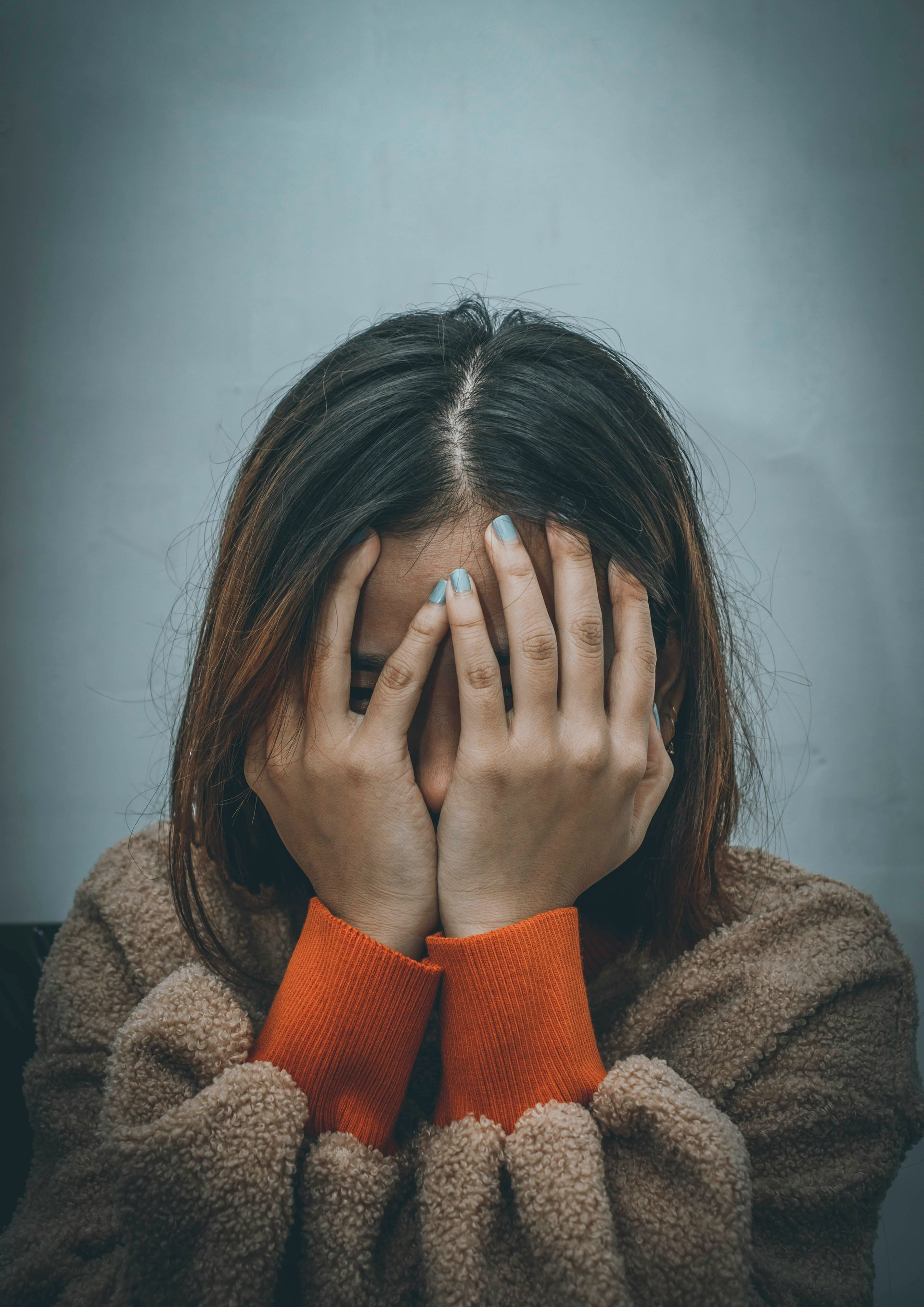 Woman covering face with hands, appearing distressed; tan sweater, orange cuffs.