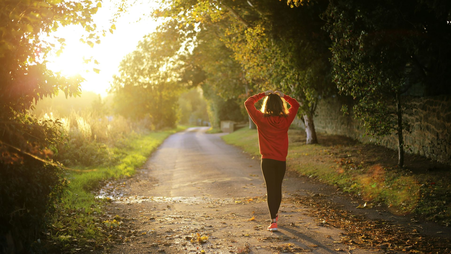 Person walking on a tree-lined path towards sunlight; wearing red jacket and black leggings.
