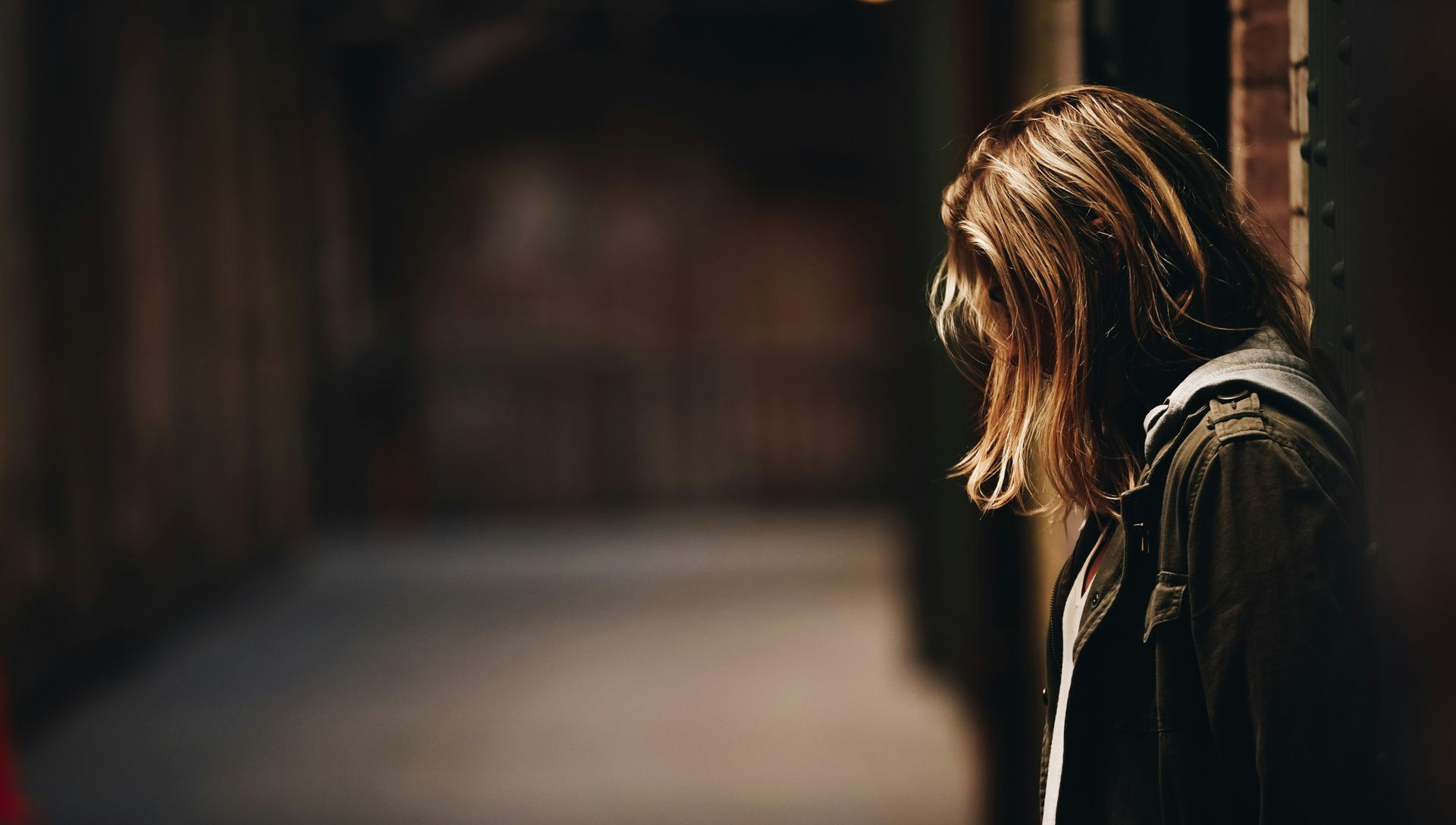 Woman in a hooded jacket stands in a dim, narrow hallway, looking down.