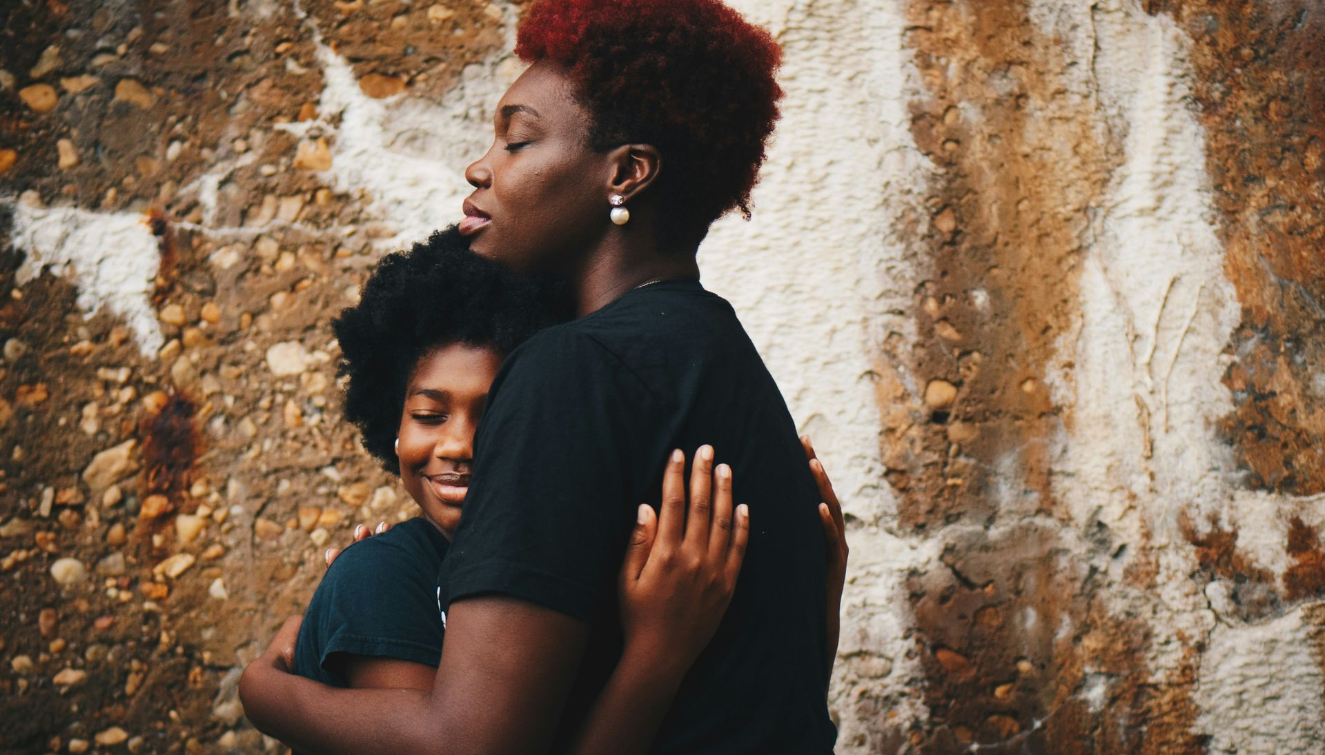 Two people embracing in front of a textured wall. One has red hair, and one has black hair. They are wearing black shirts.