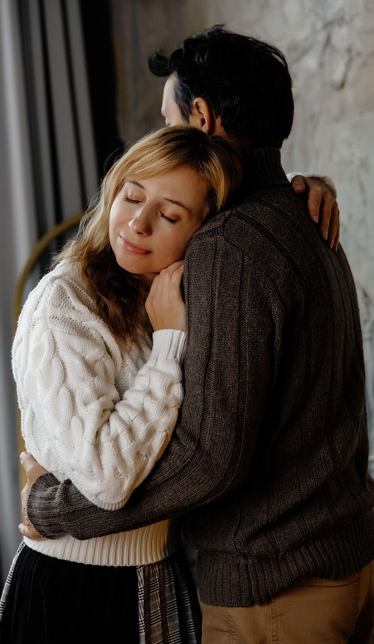 Woman in white sweater hugging a person in a brown sweater; both have eyes closed, indoor setting.