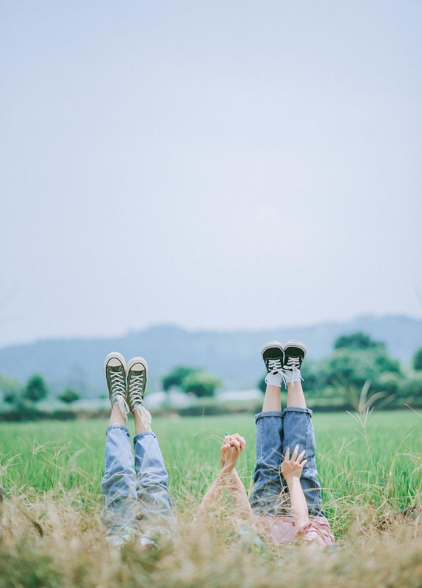 Two people lying in a grassy field with their legs raised, wearing jeans and shoes.