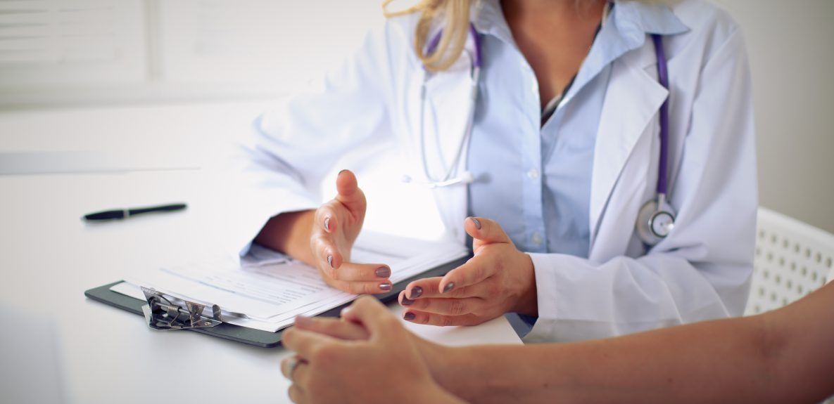 A doctor gestures while speaking to a patient in an office. A clipboard and pen sit on the table.