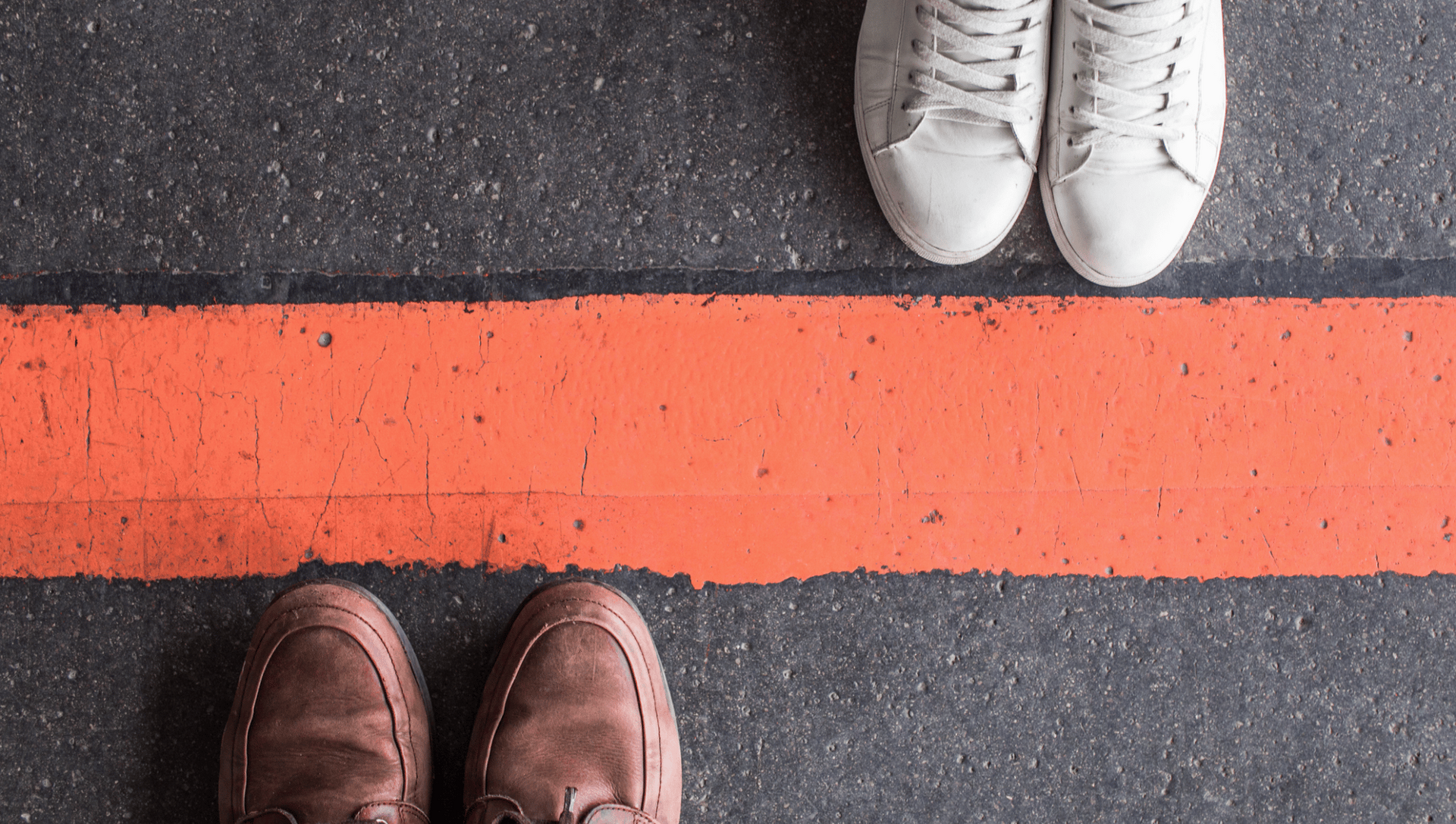 Two pairs of feet facing each other across an orange line on asphalt; one pair in brown shoes, the other in white sneakers.