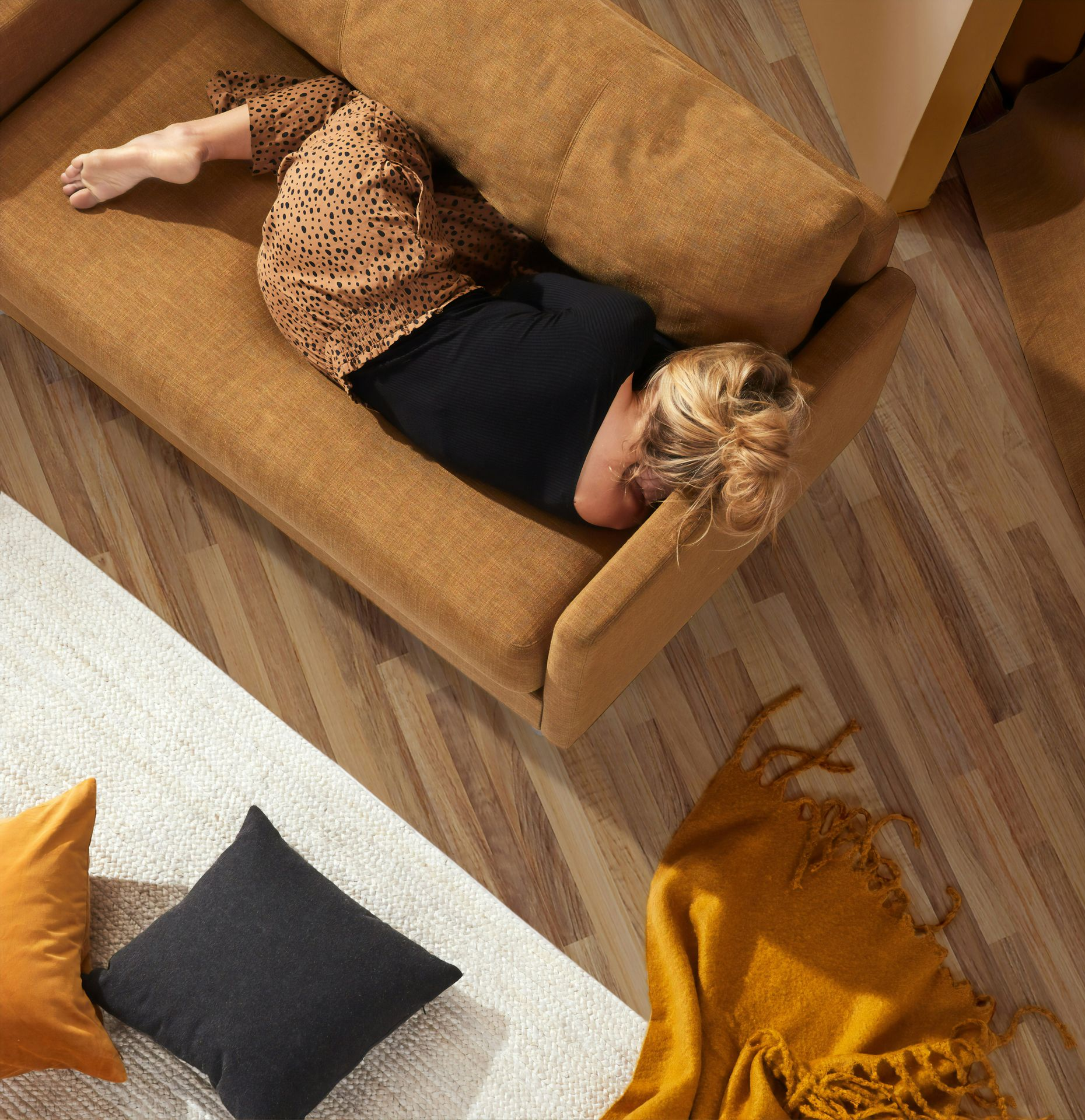 Person curled up asleep on a brown sofa, surrounded by pillows and a blanket.