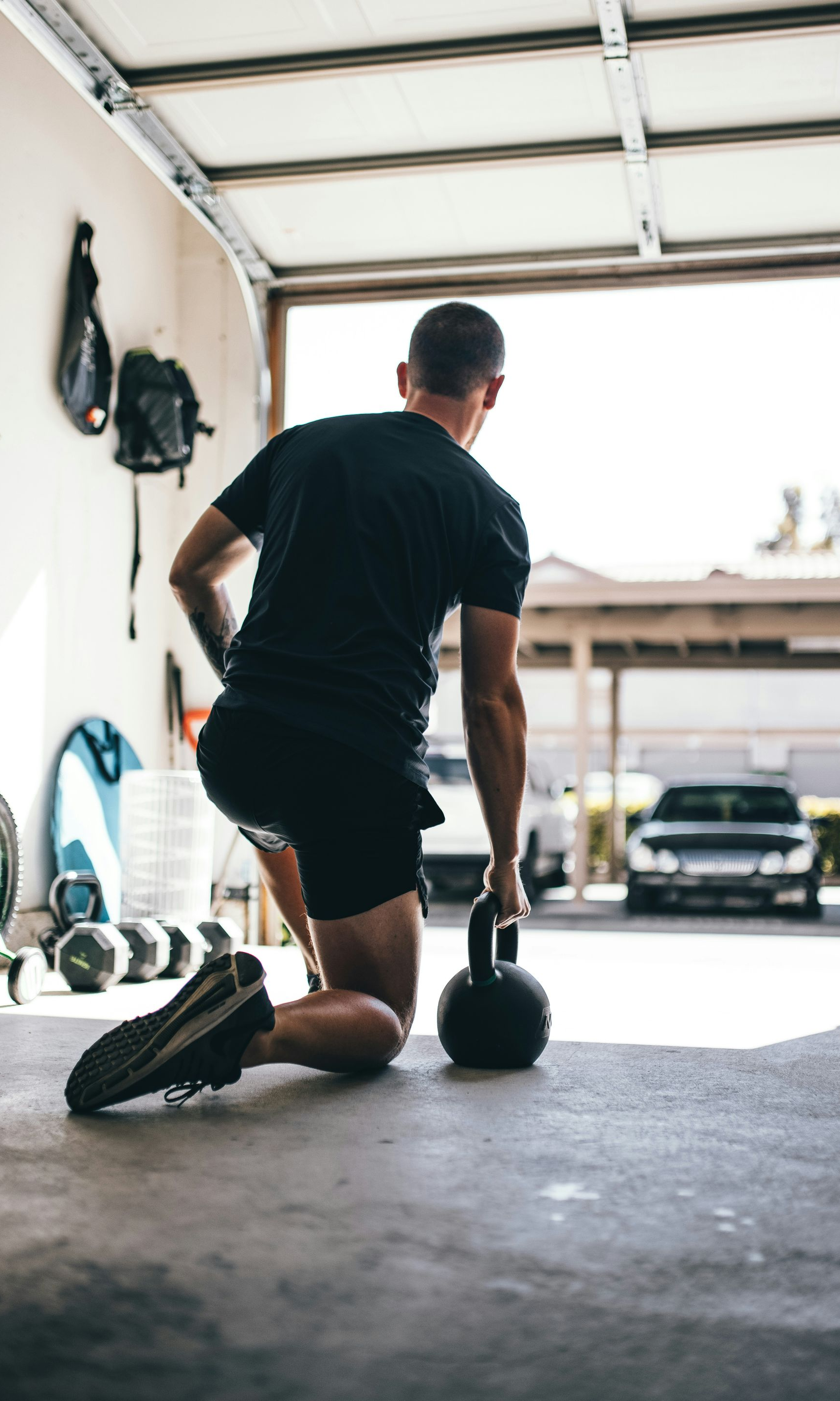 Man in black shorts and shirt lifts a kettlebell in a garage.