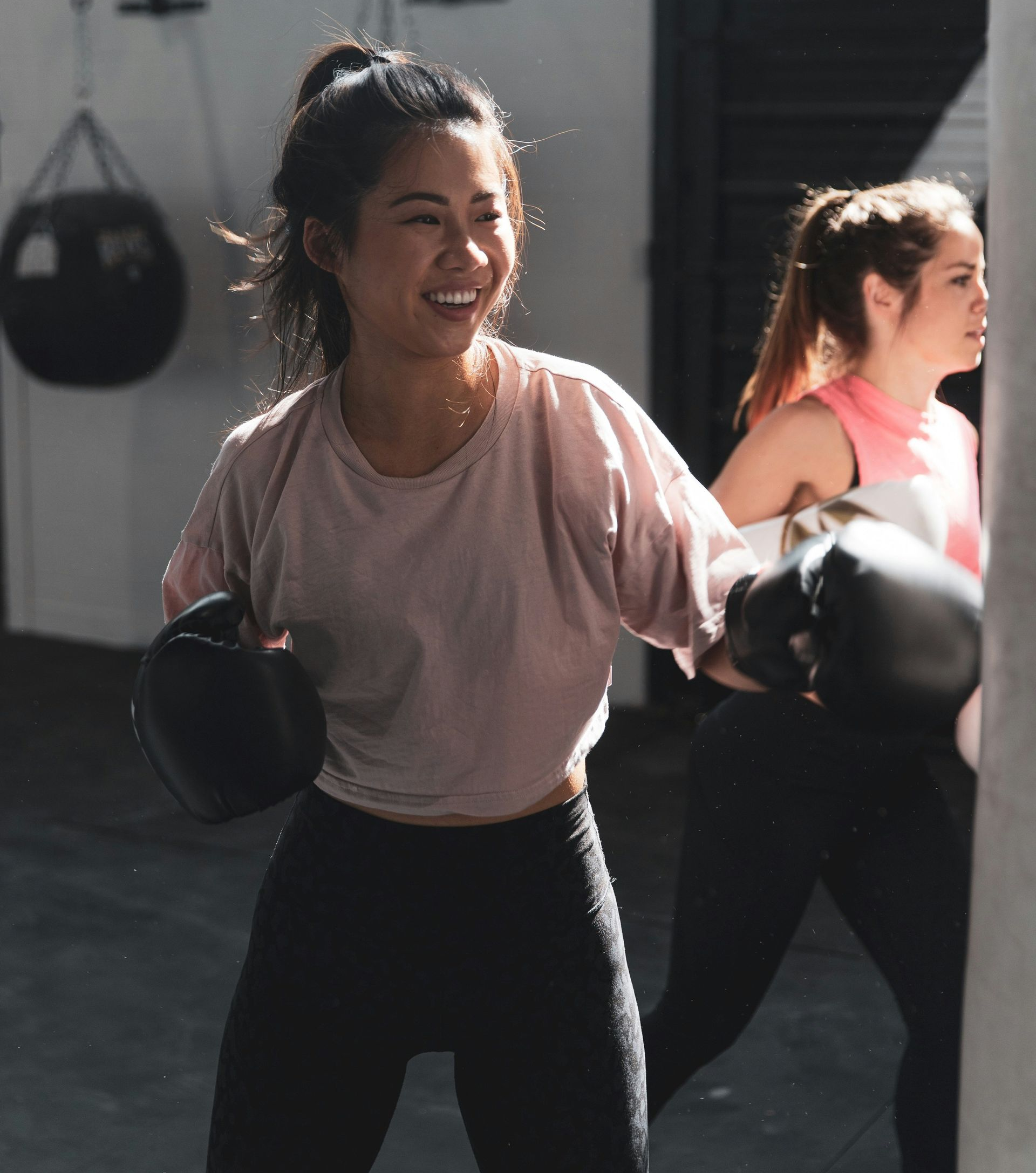 Woman in boxing gloves smiles while punching a bag in a gym; another woman in the background.