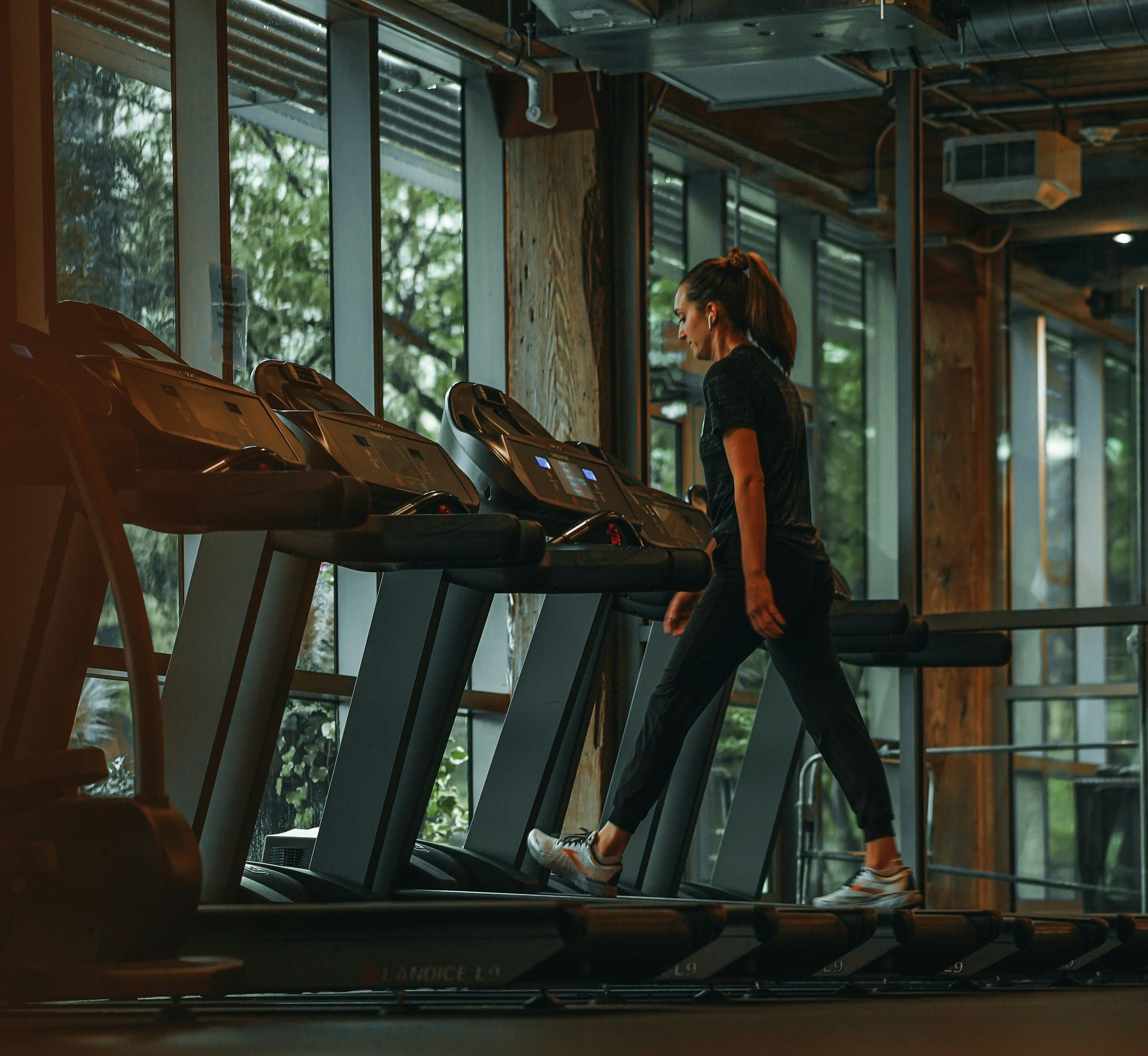 Woman in athletic wear steps onto a treadmill in a gym with large windows.