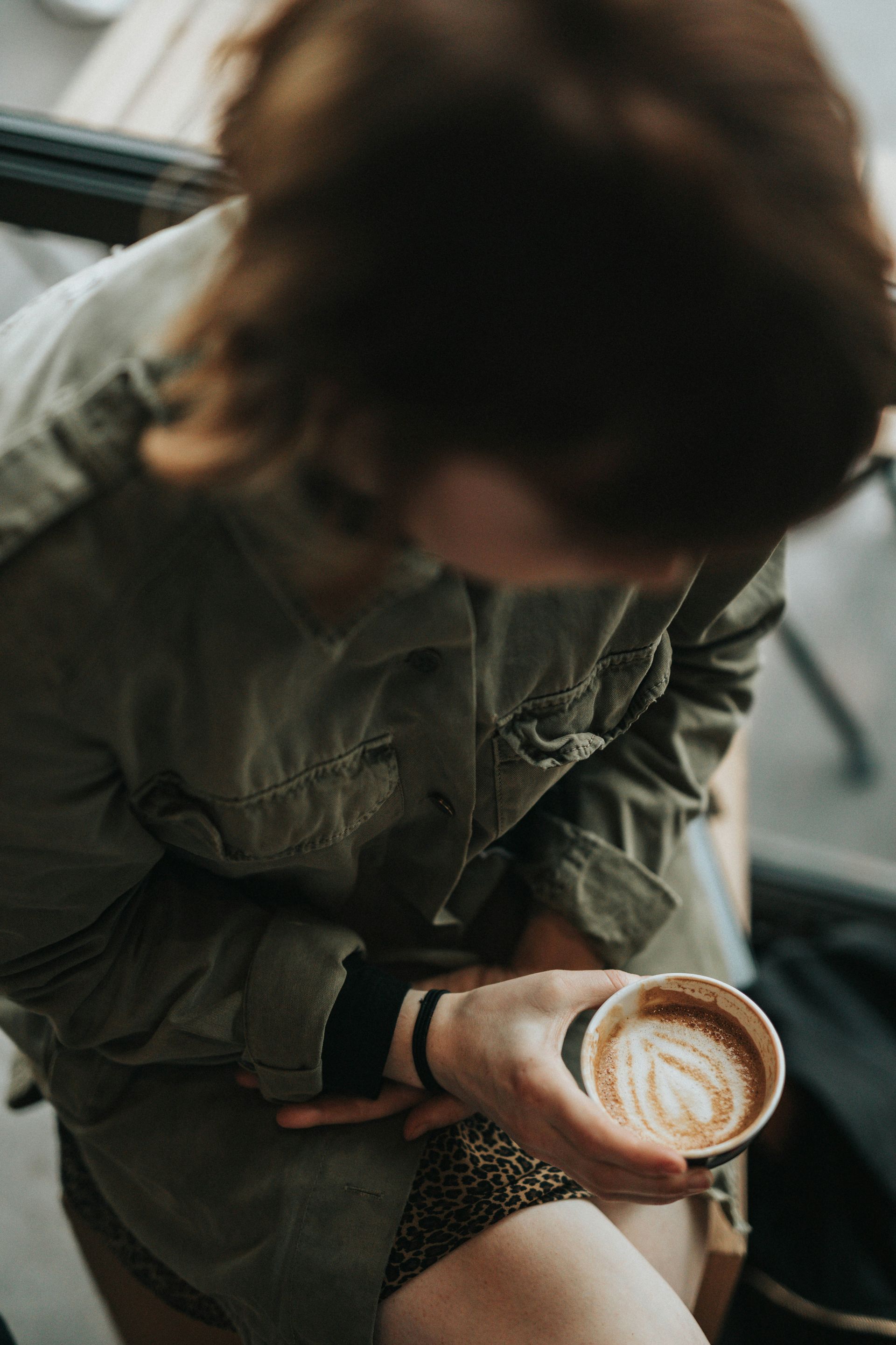 Person holding a coffee cup with latte art, wearing a green jacket, in a cafe setting.