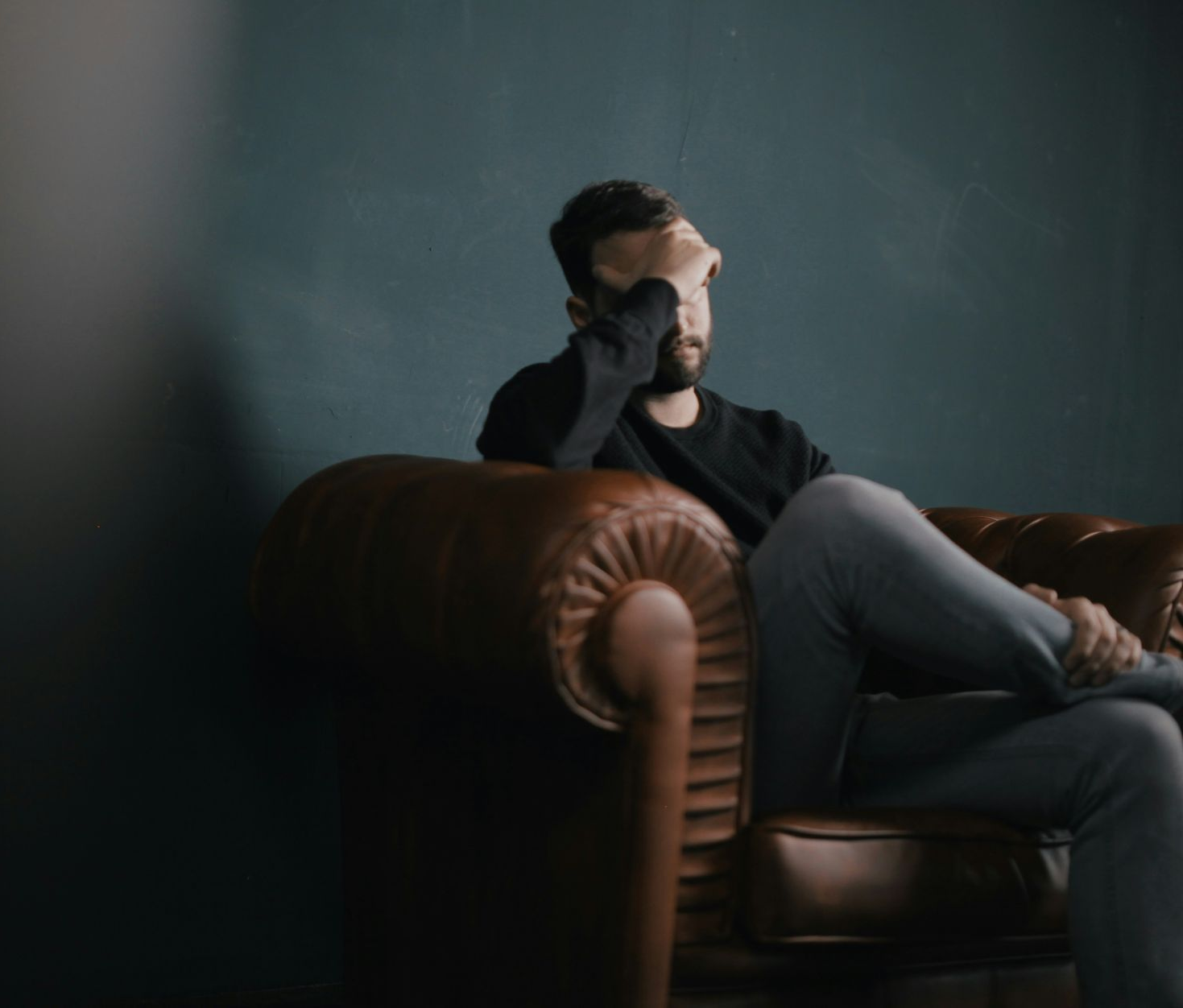 Man sits on a brown leather couch, hand on face, looking distressed. Dark blue wall in background.