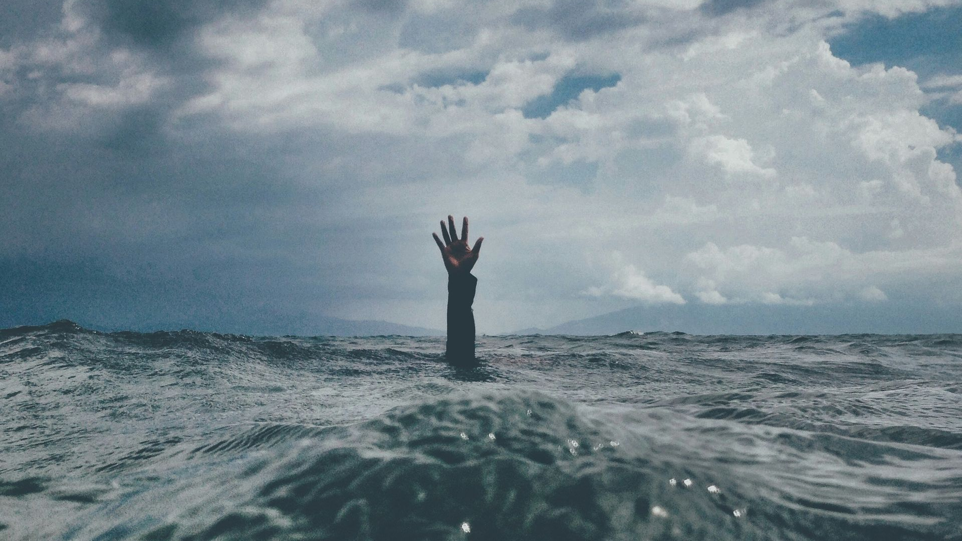 A hand reaching out of turbulent ocean waves toward a cloudy, overcast sky.