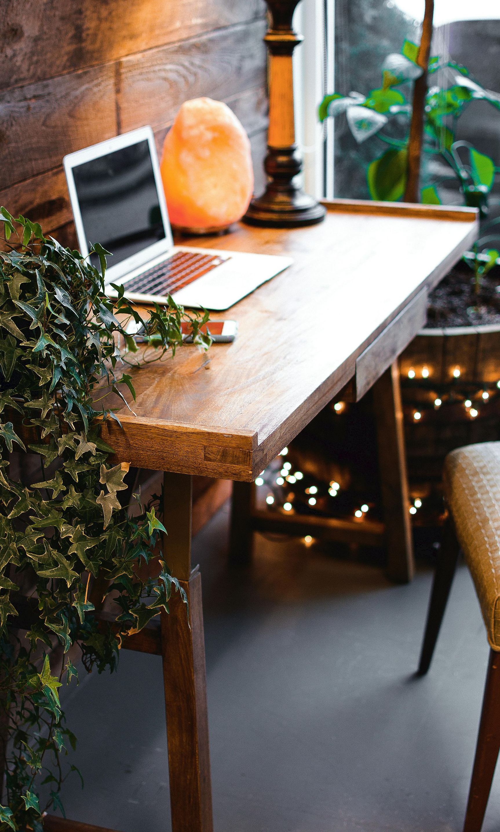Laptop on wooden desk by a window with greenery and fairy lights.