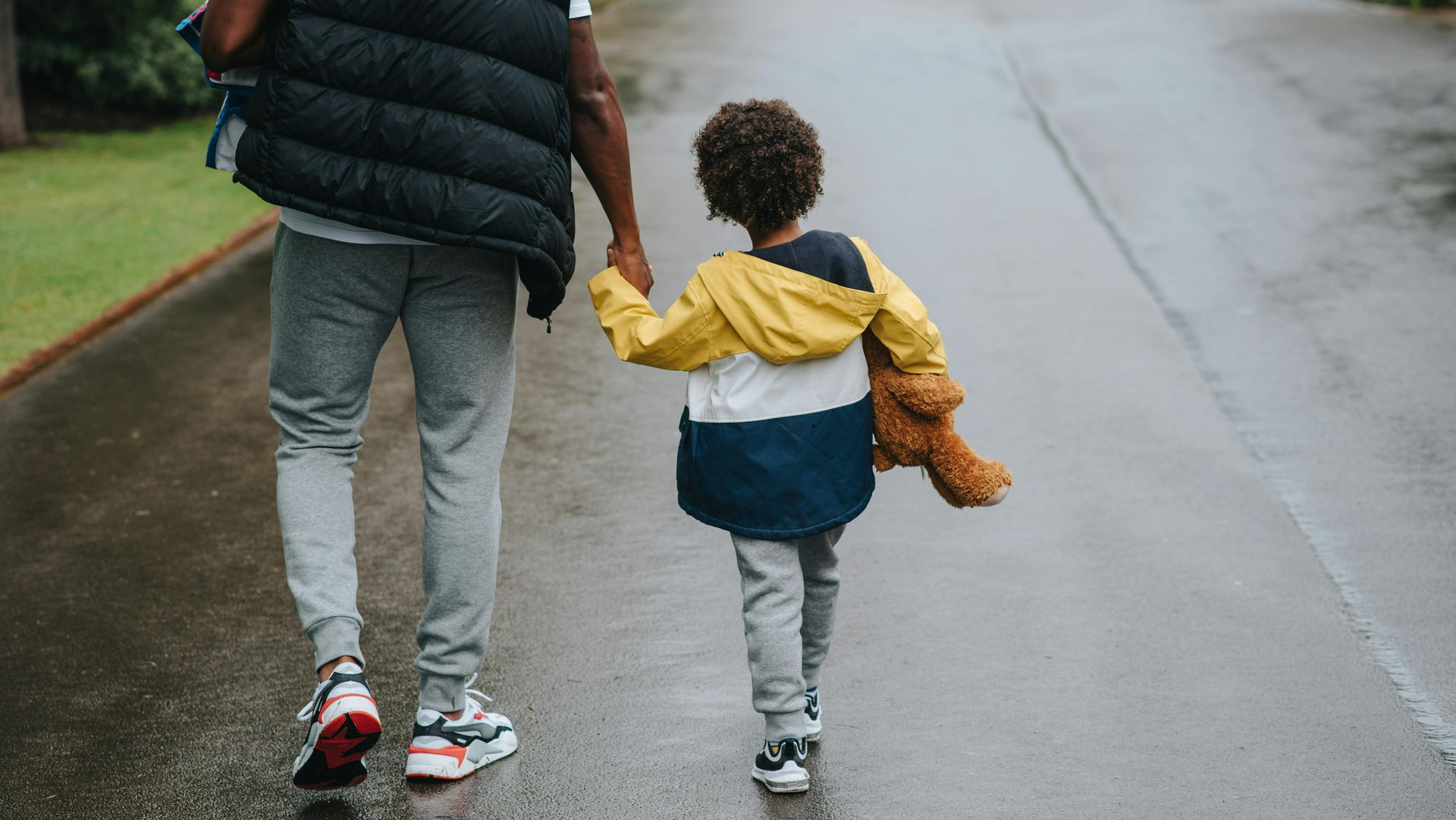 Man and child holding hands, walking on a wet path. The child carries a teddy bear.