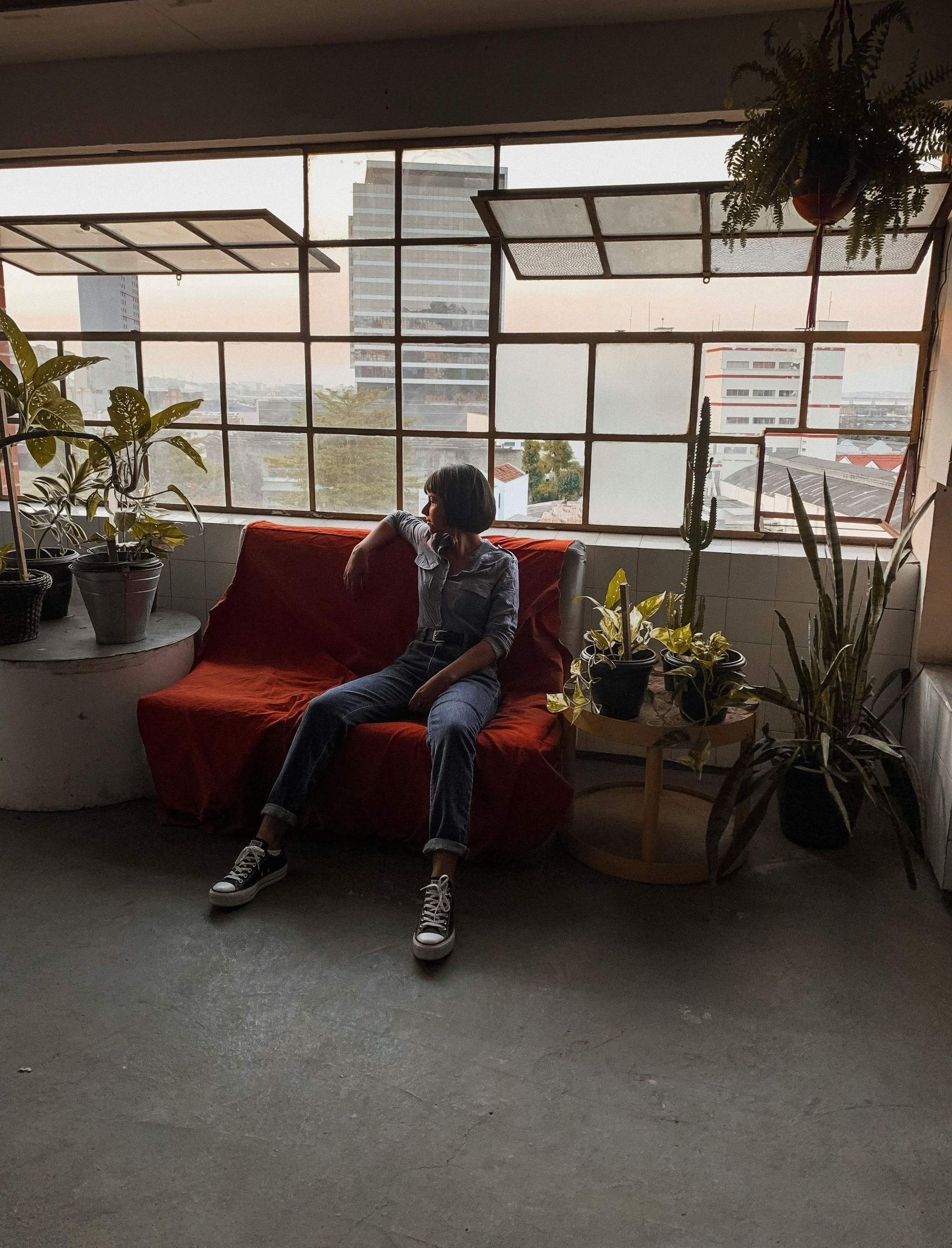 Person sitting on a red sofa by a large window with city views, surrounded by plants.
