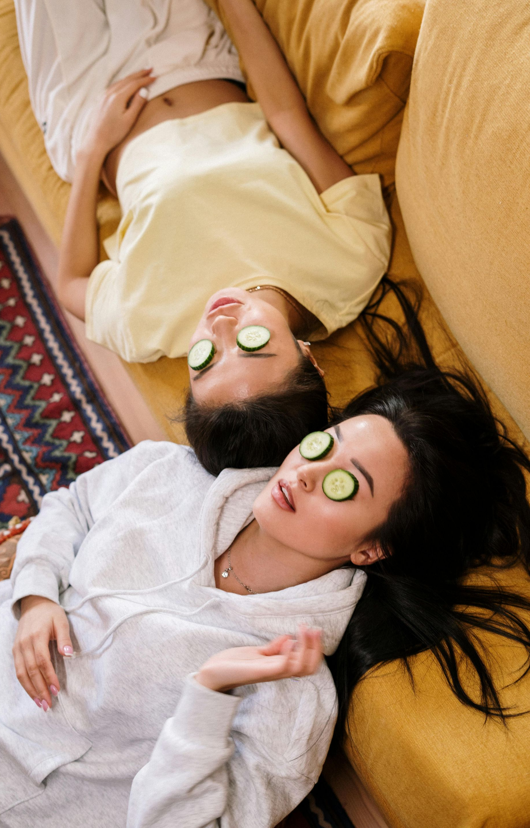Two people relaxing on a yellow couch with cucumber slices on their eyes.