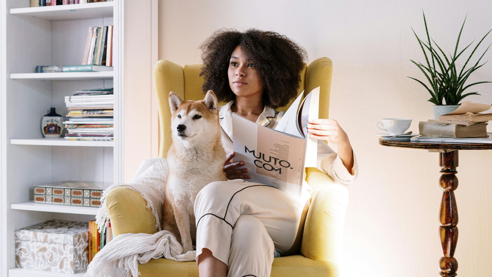Woman with dog in yellow armchair reading a magazine near a bookshelf and table.