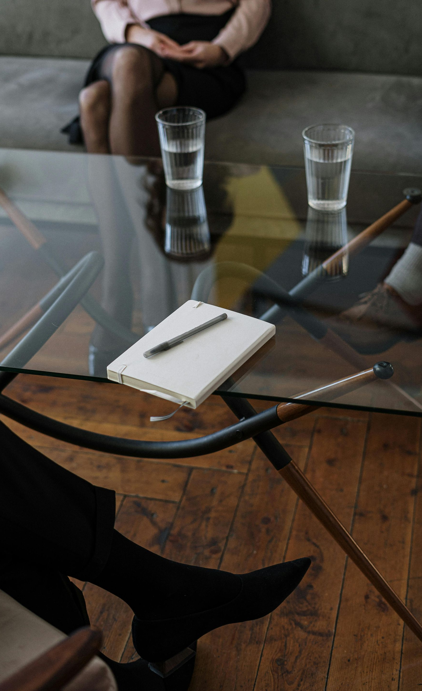 Glass table with notepad, two glasses of water, and legs of people seated in a therapy session.