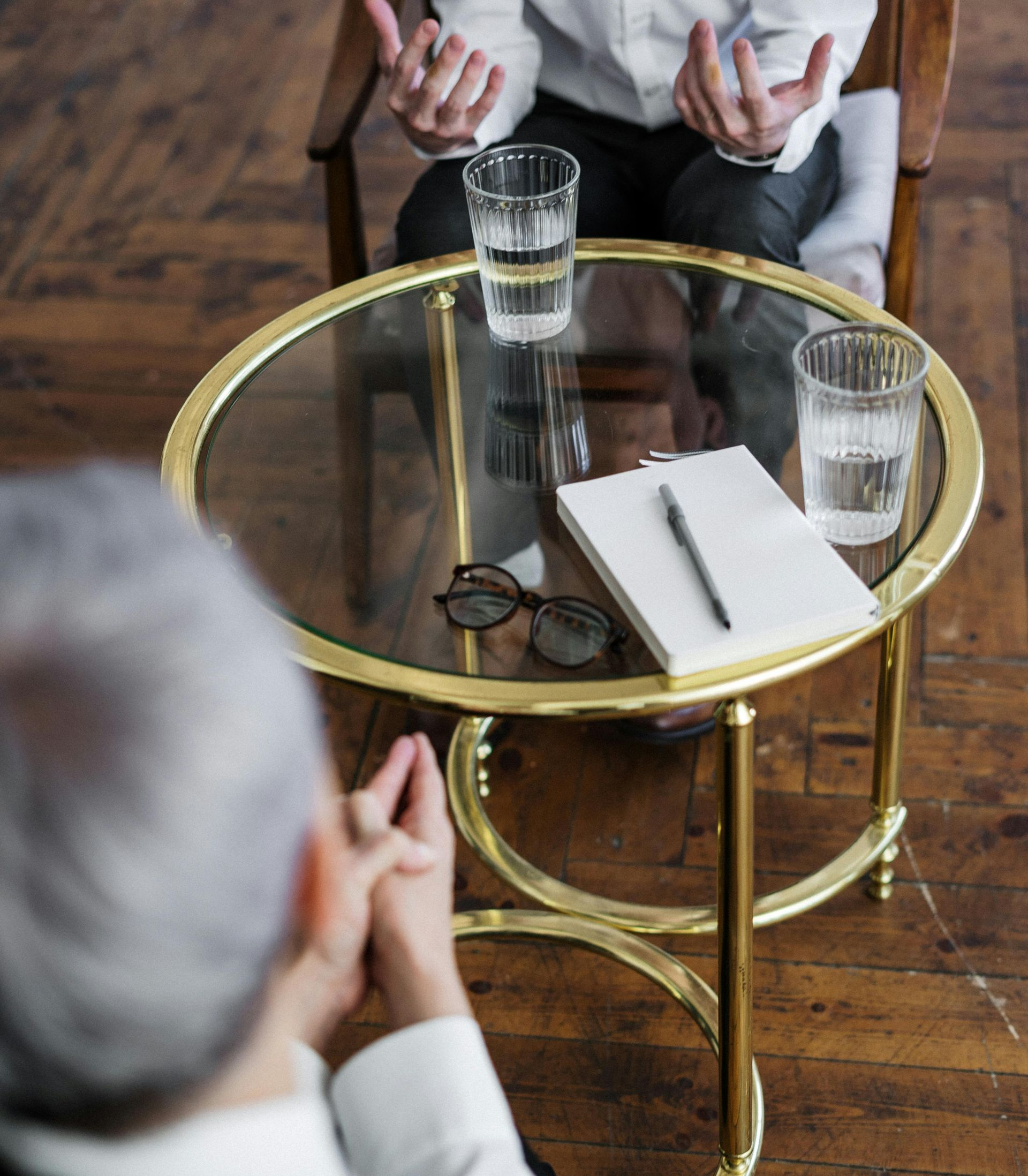 Two people in a therapy session. One gestures while the other listens. Table holds water, notepad, and glasses.