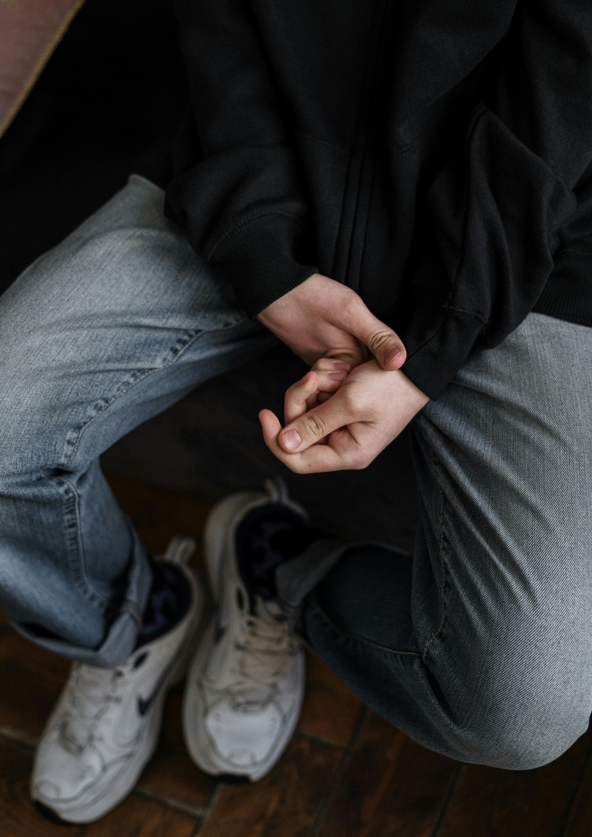 Person in black sweater and jeans, hands clasped in lap. Worn white sneakers.