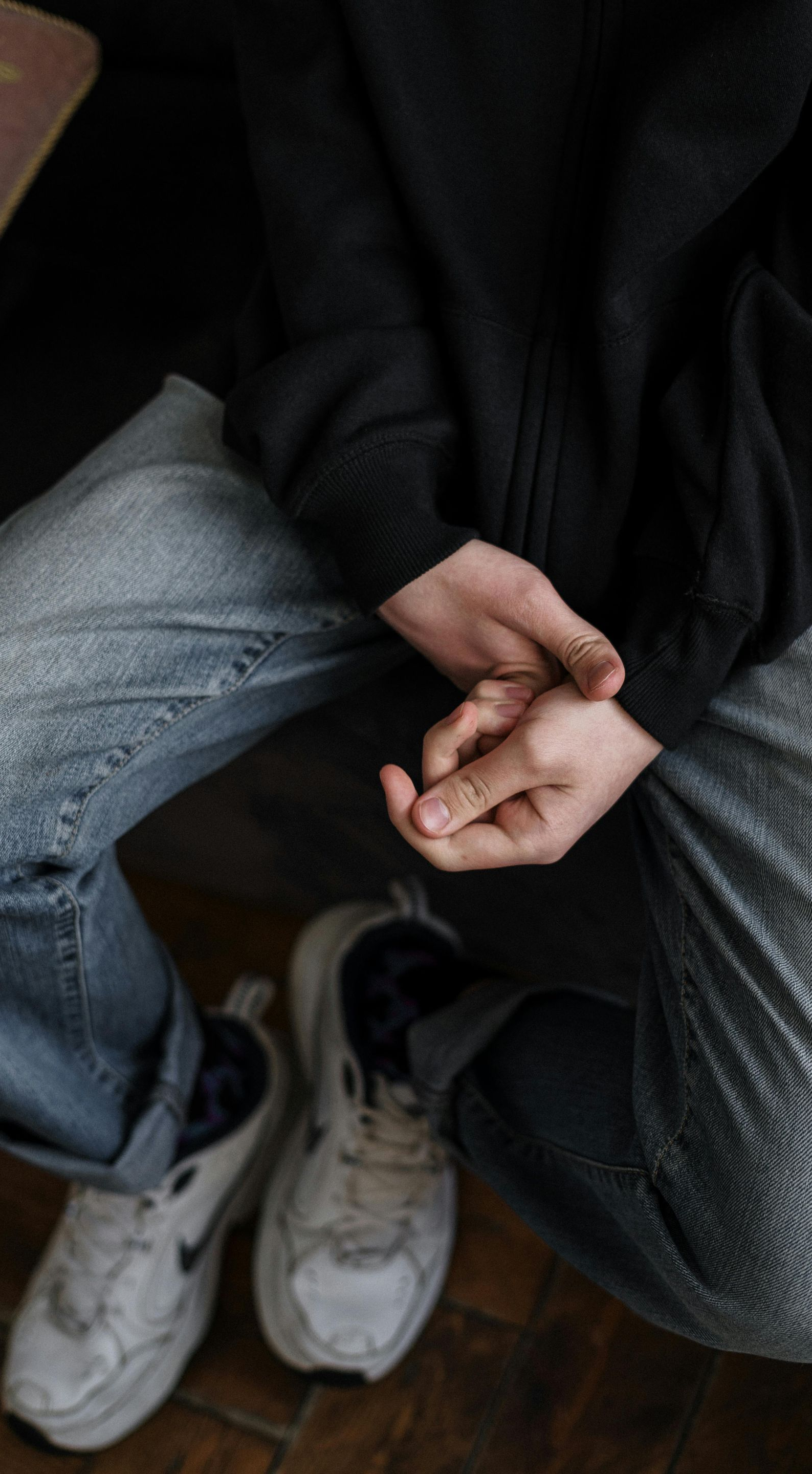 Person in black sweater and jeans, hands clasped in lap. Worn white sneakers.