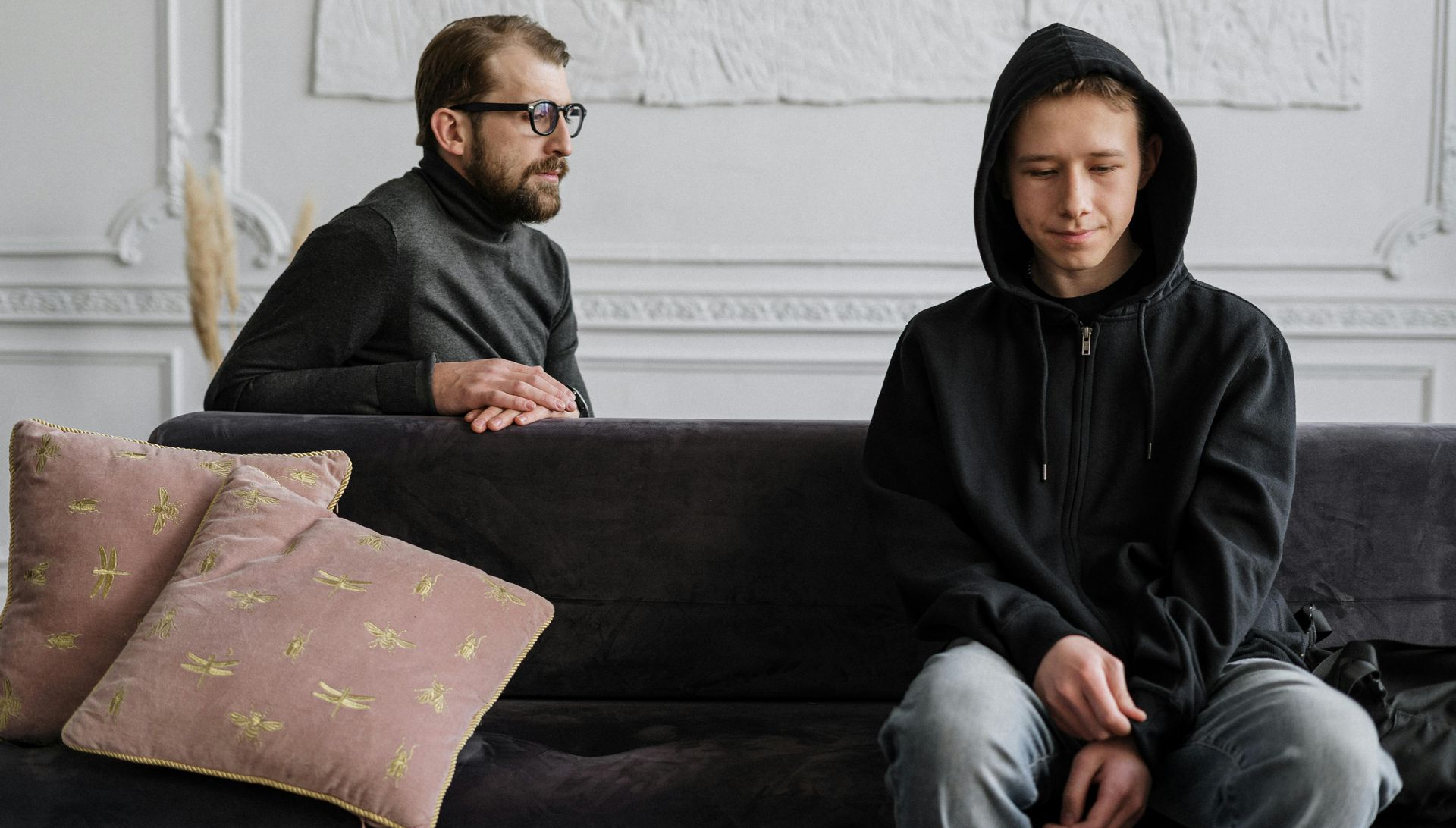Man in glasses watches teen in black hoodie sit on a sofa.