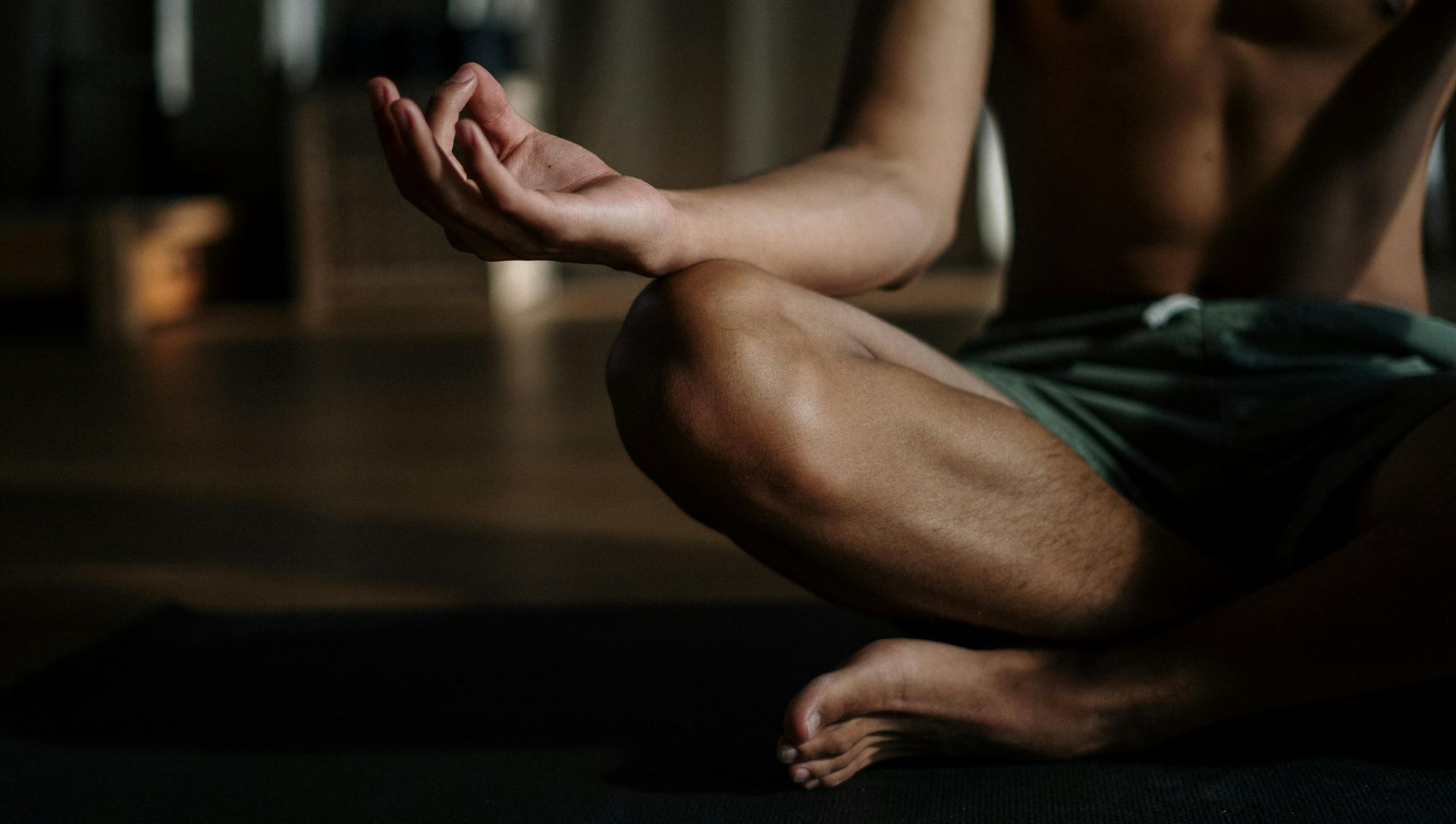 Man meditating cross-legged, torso bare, hands in mudra. Dark room, sunlight, yoga mat.