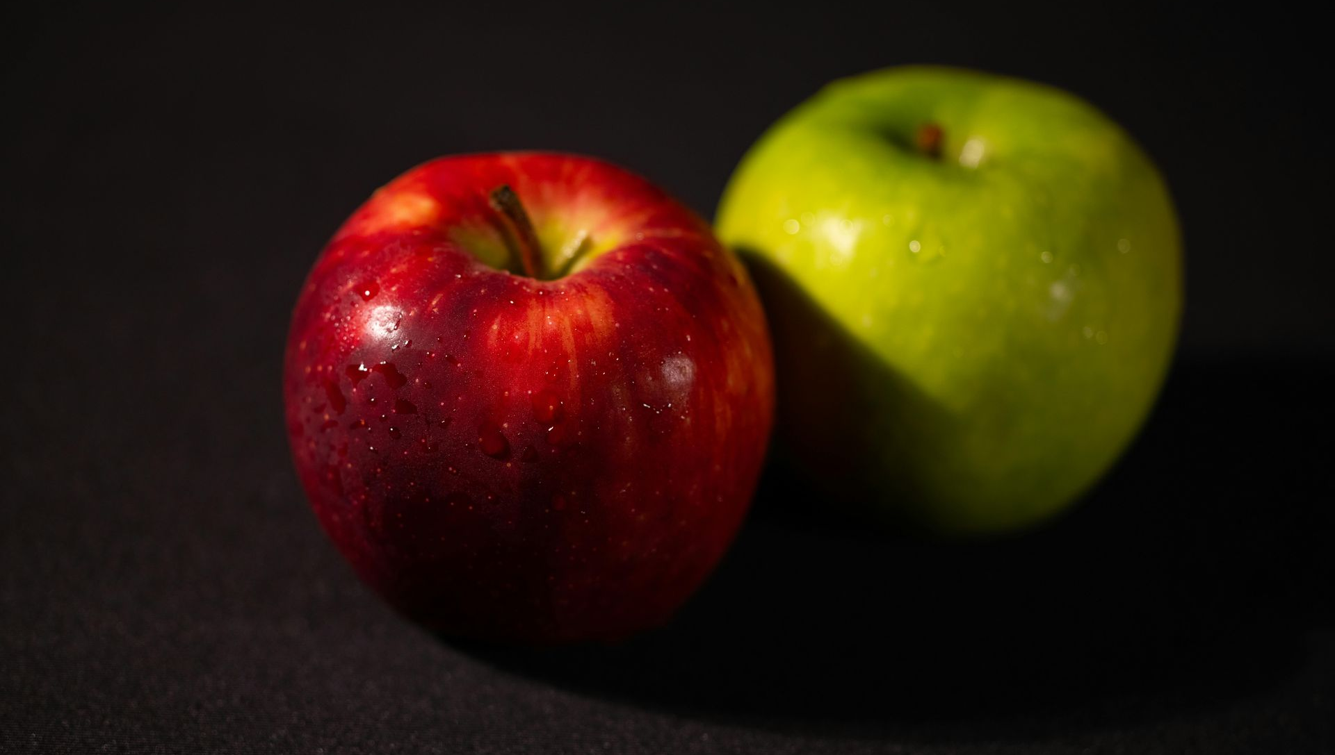 Red and green apples on a dark surface, lit from above, showing details in the fruit's skin.
