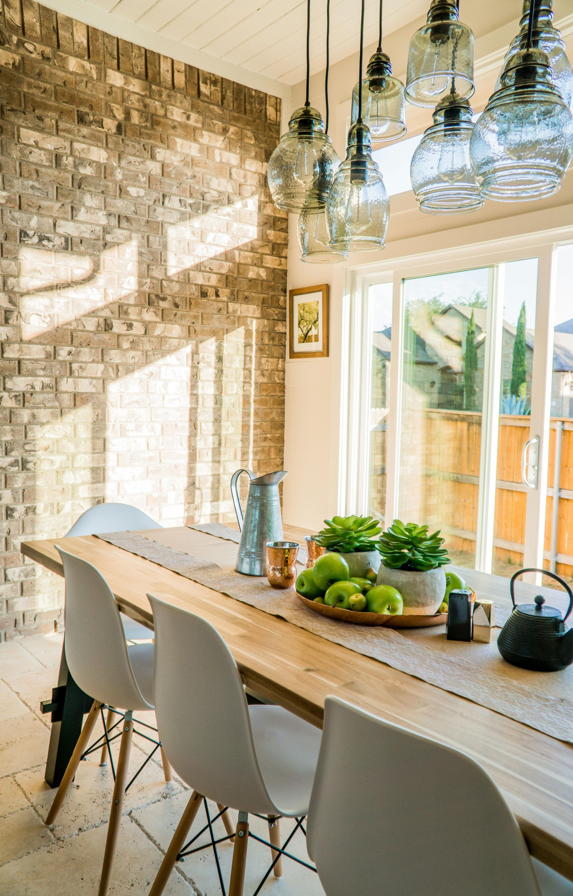Dining room with long wooden table, white chairs, brick wall, glass pendant lights, and sliding glass door.