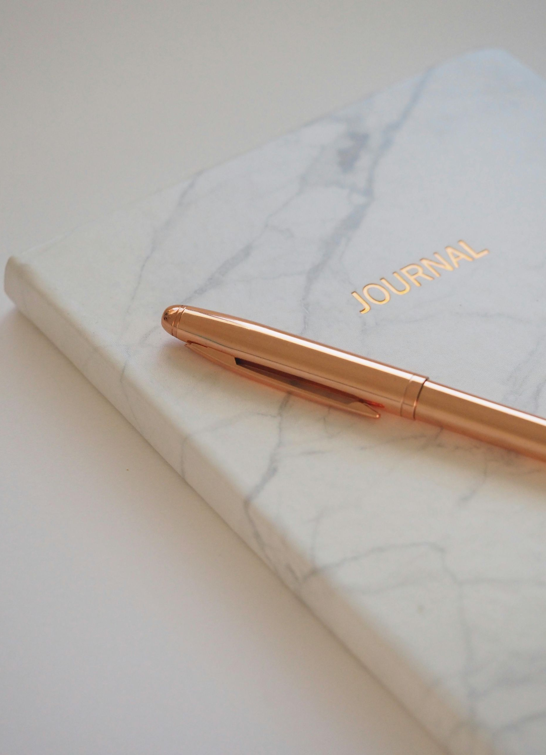 Marble journal with gold lettering, rose-gold pen on white surface.