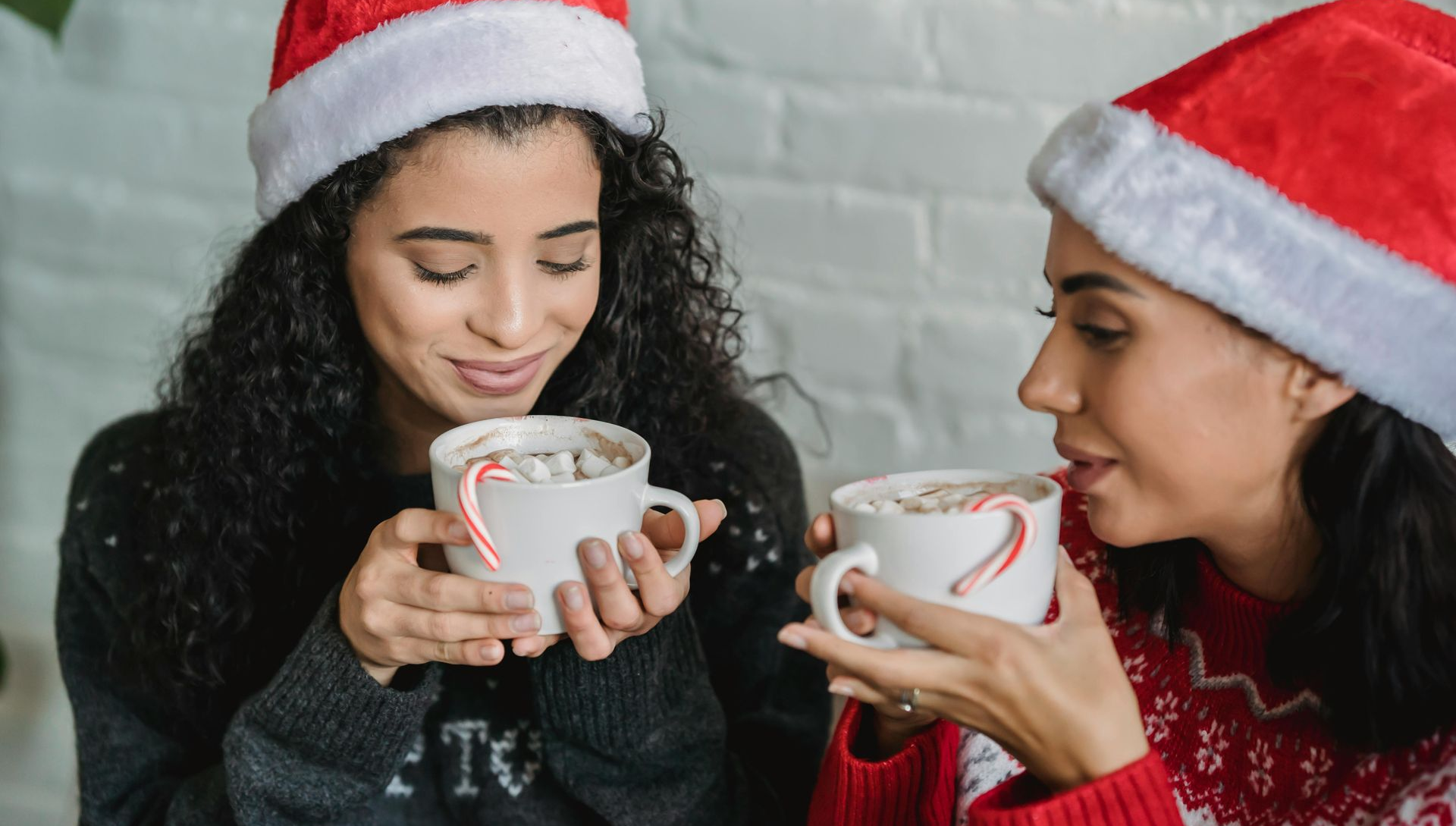 Two people in Santa hats sip hot cocoa with candy canes.