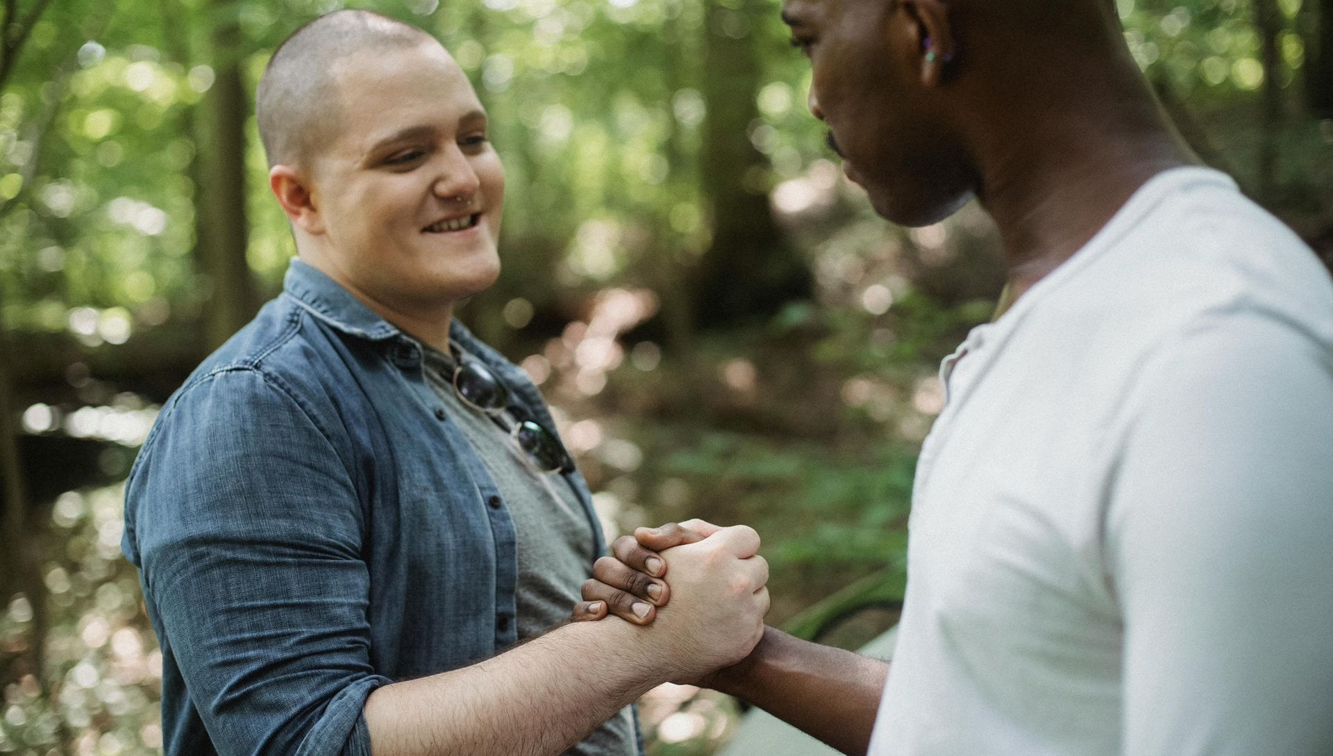 Two people shaking hands outdoors in a forest. One with a shaved head smiles, wearing denim. The other wears a white shirt.