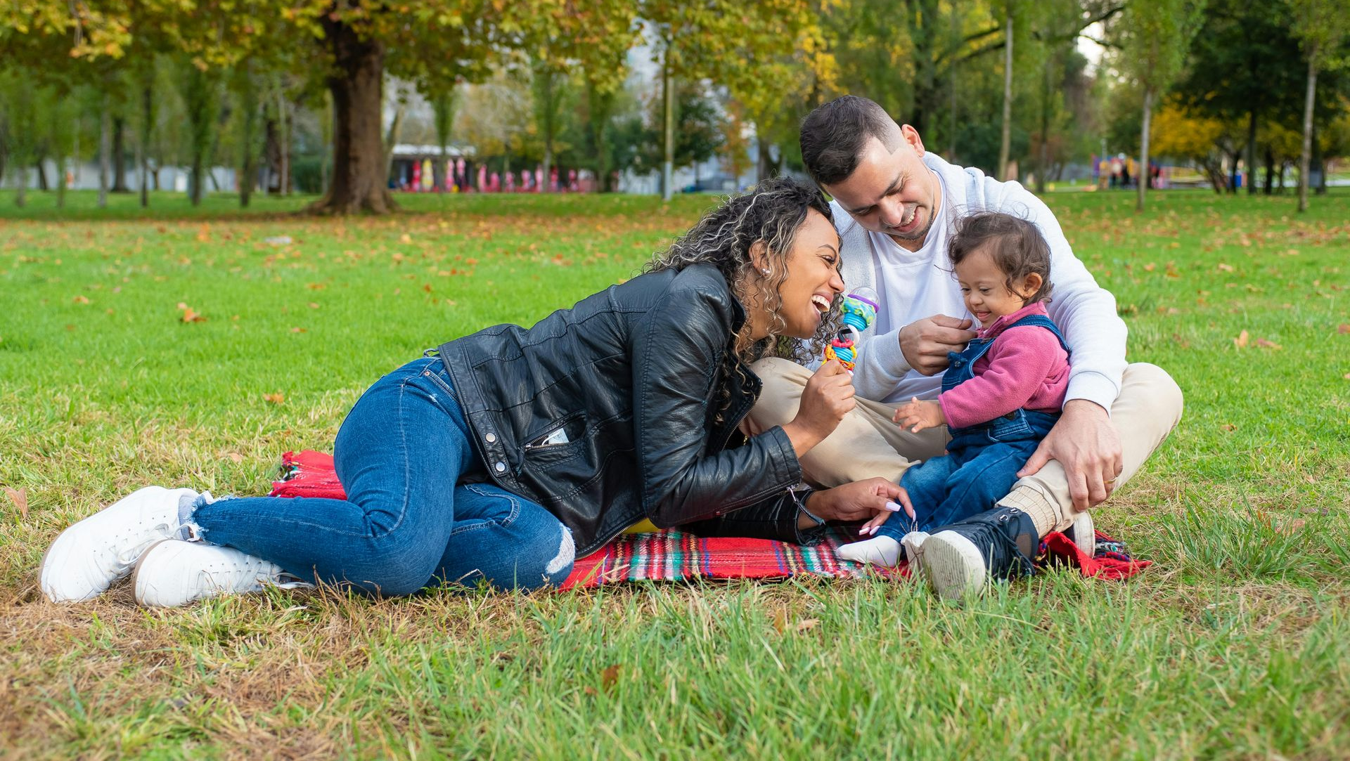 Family on a picnic blanket in a park, laughing and playing with a toddler.